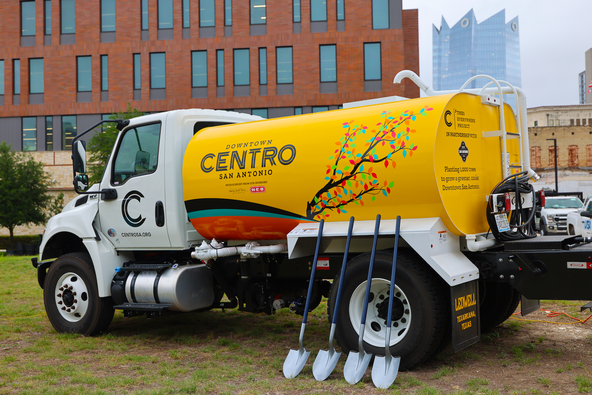 White truck with a yellow tanker, Downtown Centro San Antonio, sits on grass. A colorful tree graphic decorates the tank; shovels lean by the rear wheel; brick and glass buildings in background.