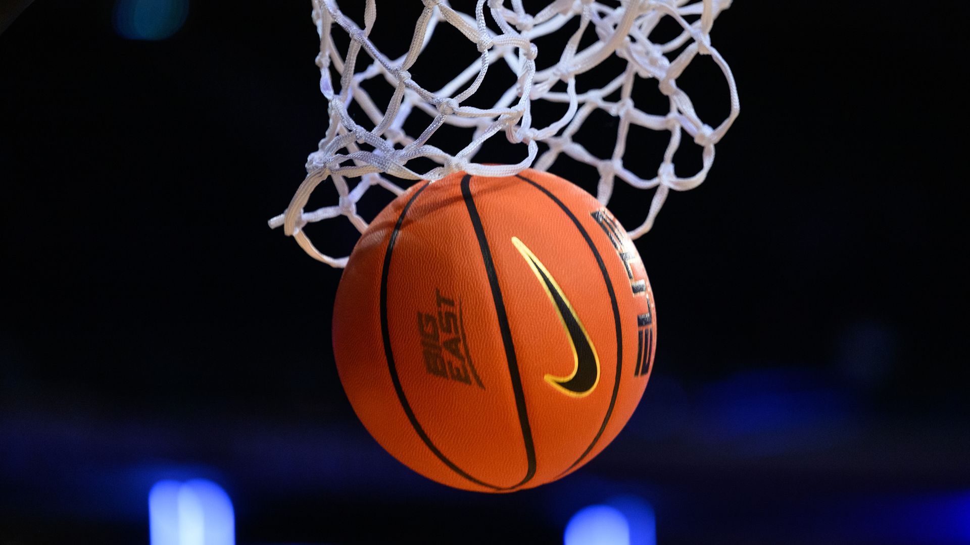Close-up of an orange basketball with a yellow Nike swoosh hanging in a white basketball net, against a dark arena with blue lights in the background.
