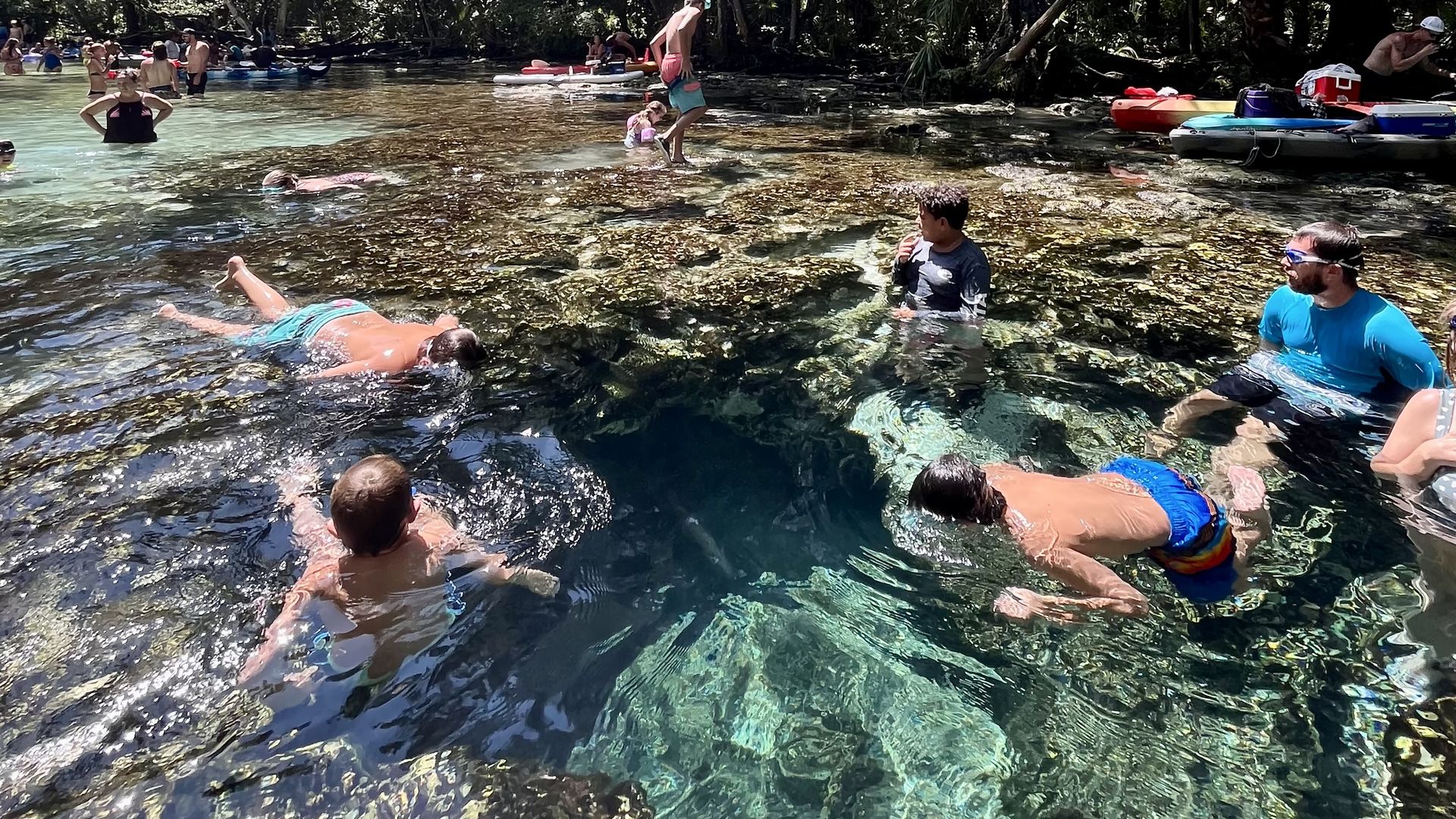 Clear, turquoise water reveals a deep spring surrounded by rocks. Two boys in blue swim trunks swim at the opening of the spring, surrounded by kayaks and other swimmers.