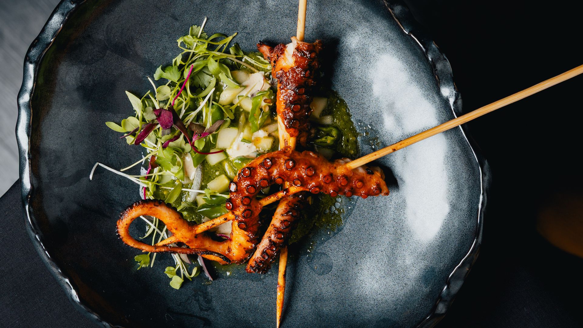 Top-down view of a dark ceramic plate with grilled octopus skewers resting on a bed of greens and diced vegetables, set against a black background.