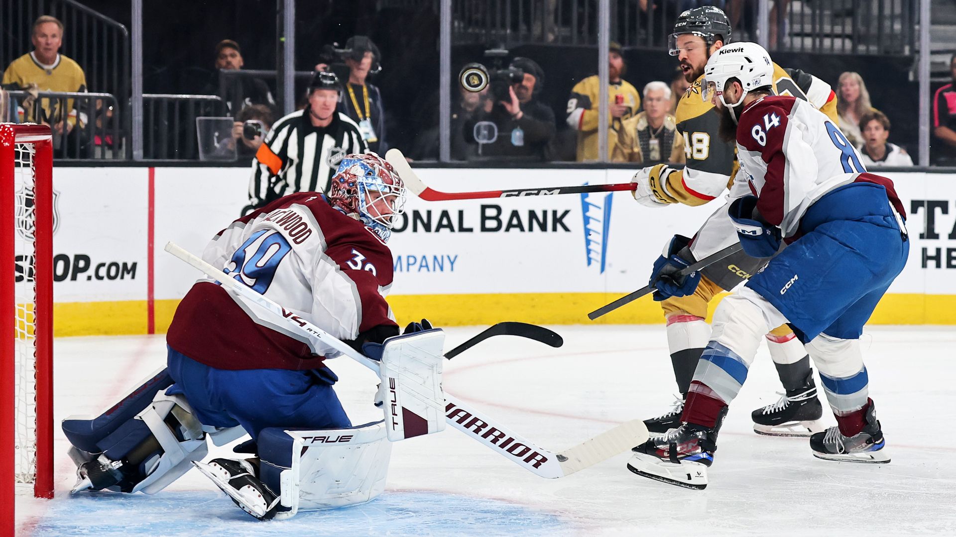 Ice hockey scene: a goalie in maroon and white guards the net while two players, one in maroon/white and one in gold, battle for a puck midair; crowd and referee in background.