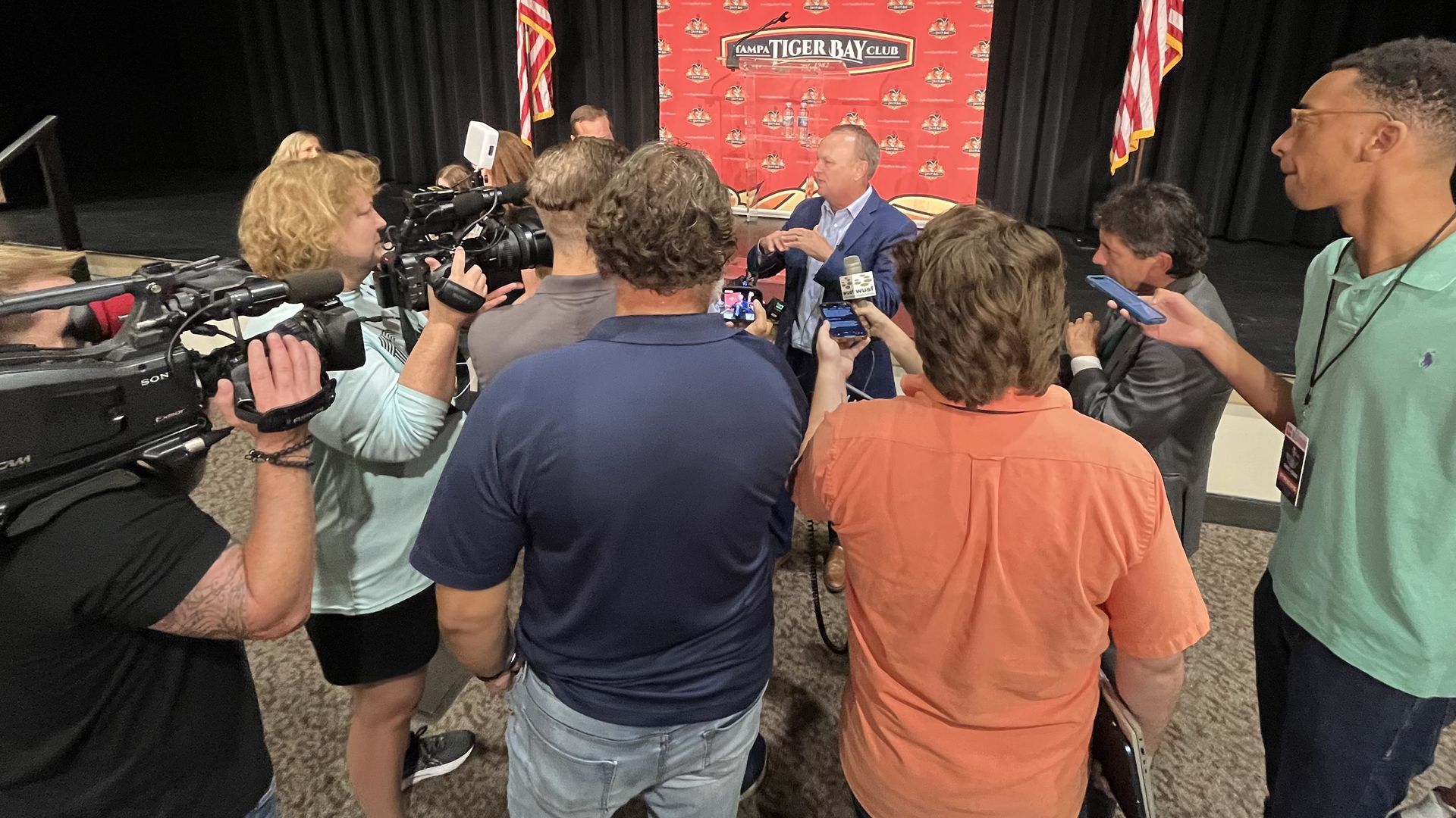 A man surrounded by reporters holding microphones and cameras. In the background, a red poster says "Tampa Tiger Bay Club."