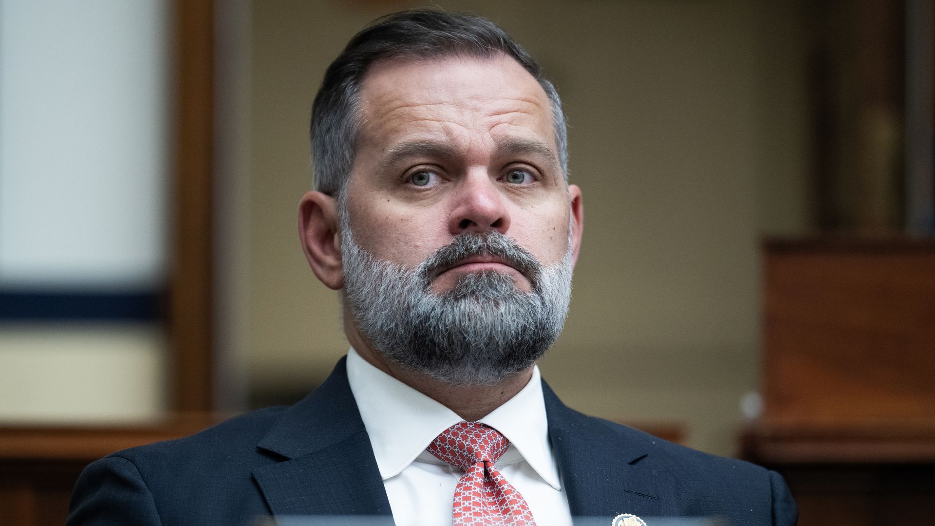 Close-up of a middle-aged man with a gray beard, wearing a dark suit, white shirt, and a red patterned tie, seated in a formal setting with wooden panels in the background.