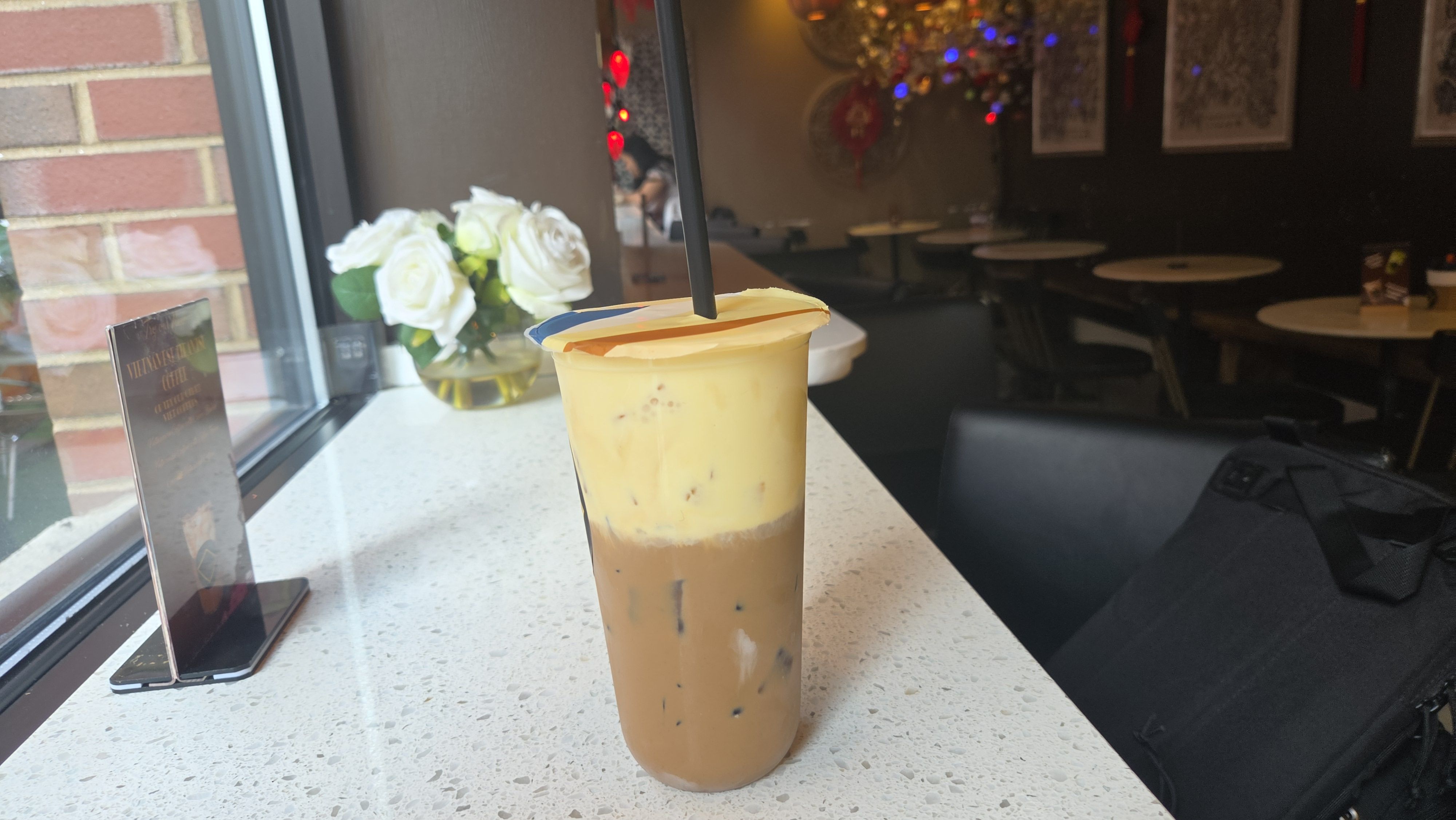 Tall iced latte in a clear cup with a black straw on a speckled white counter by a window; white roses in a vase nearby, and a dim cafe with round tables and framed art in the background.