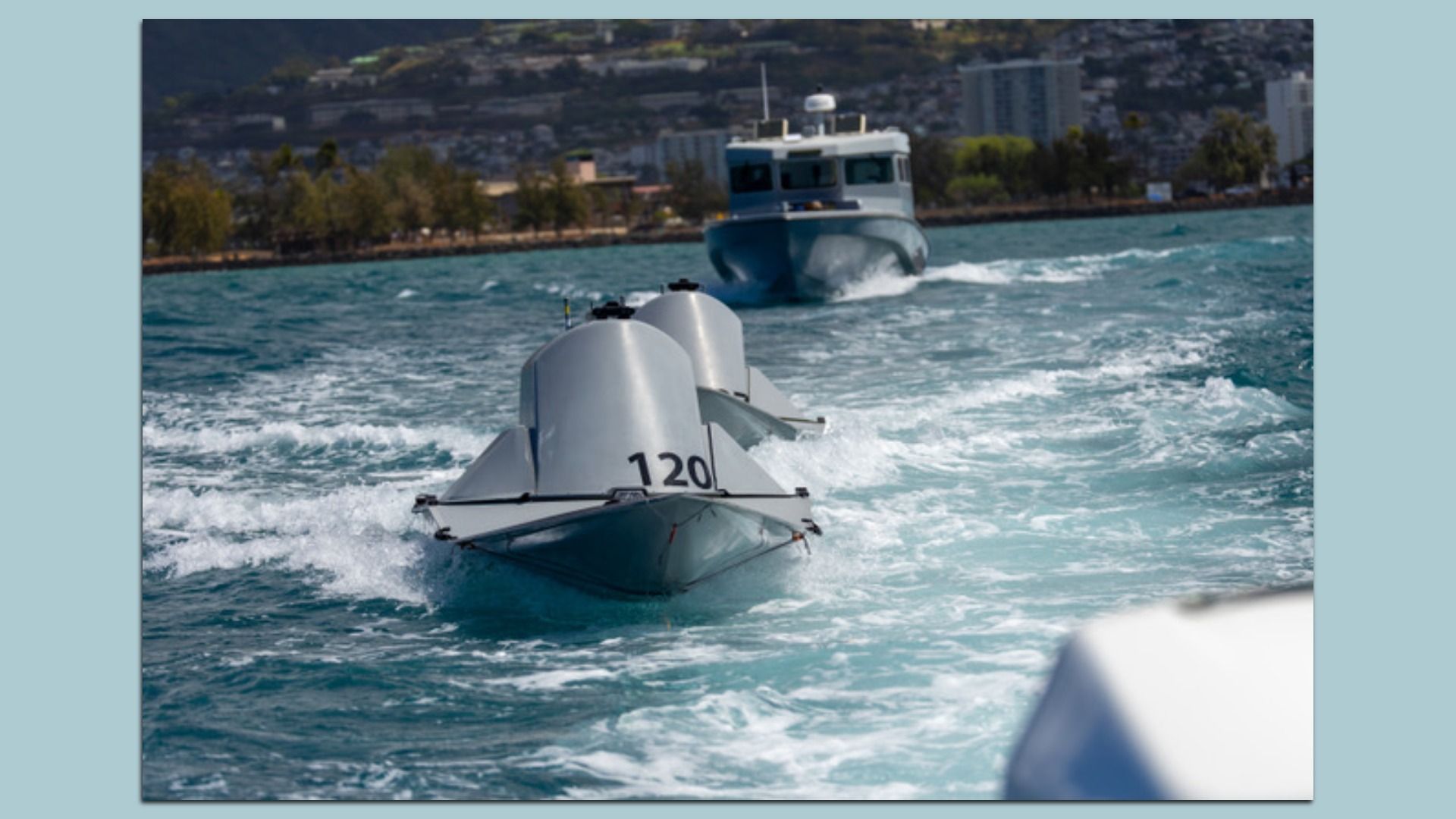 Three gray autonomous boats moving through choppy blue water near a shoreline with trees and buildings.