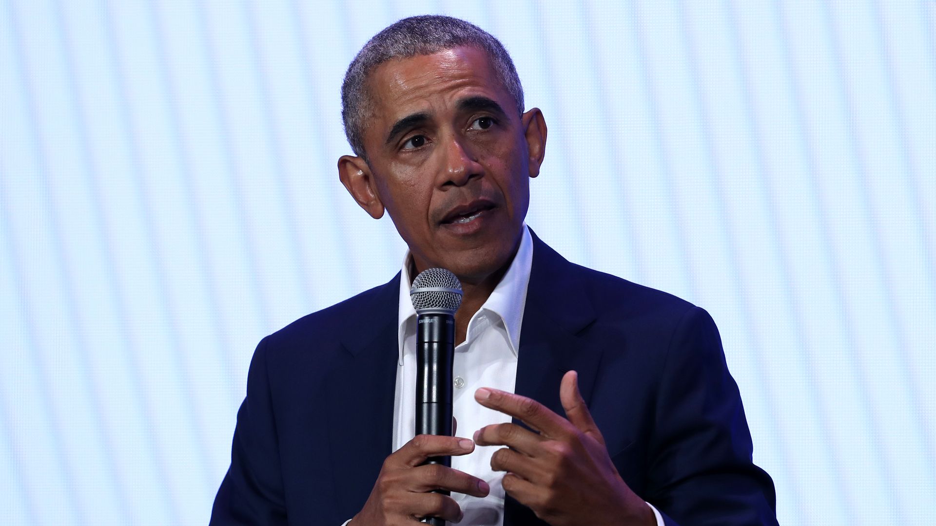 In this image, former president Barack Obama sits in front of a light blue background speaking into a microphone.