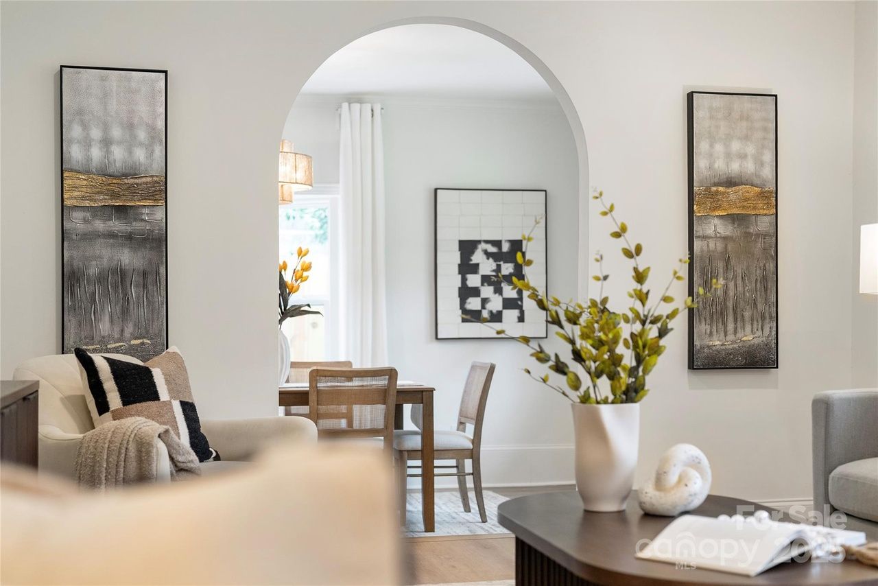 Modern living room with neutral tones, beige armchair with striped pillow, abstract wall art with gold accents, an archway leading to a dining area with wooden table and chairs, and green plant on coffee table.