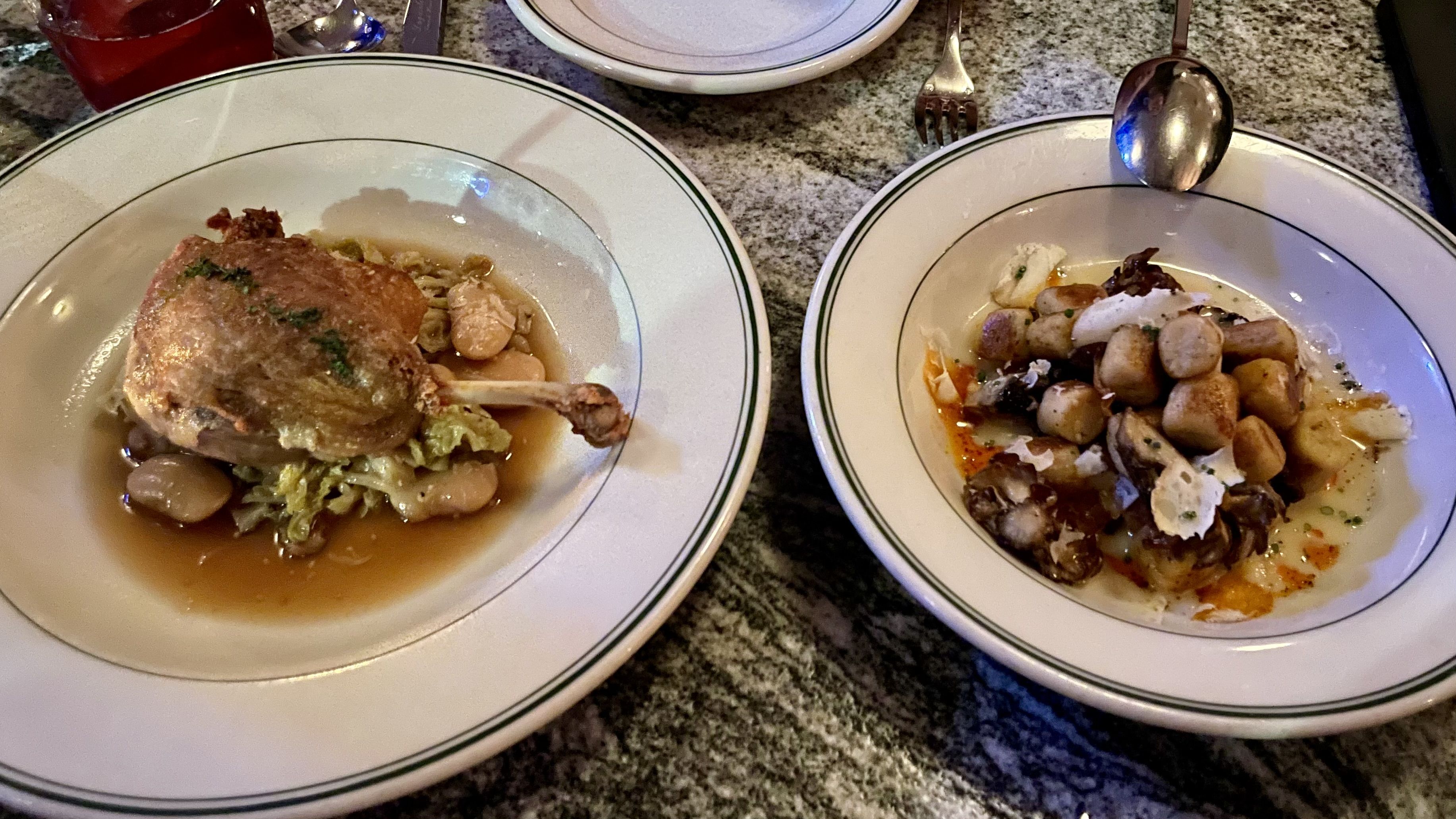 Two white plates with green rims on a granite table. Left shows a roasted quail atop cabbage and beans in brown sauce; right features cubed potatoes with mushrooms in a creamy sauce.