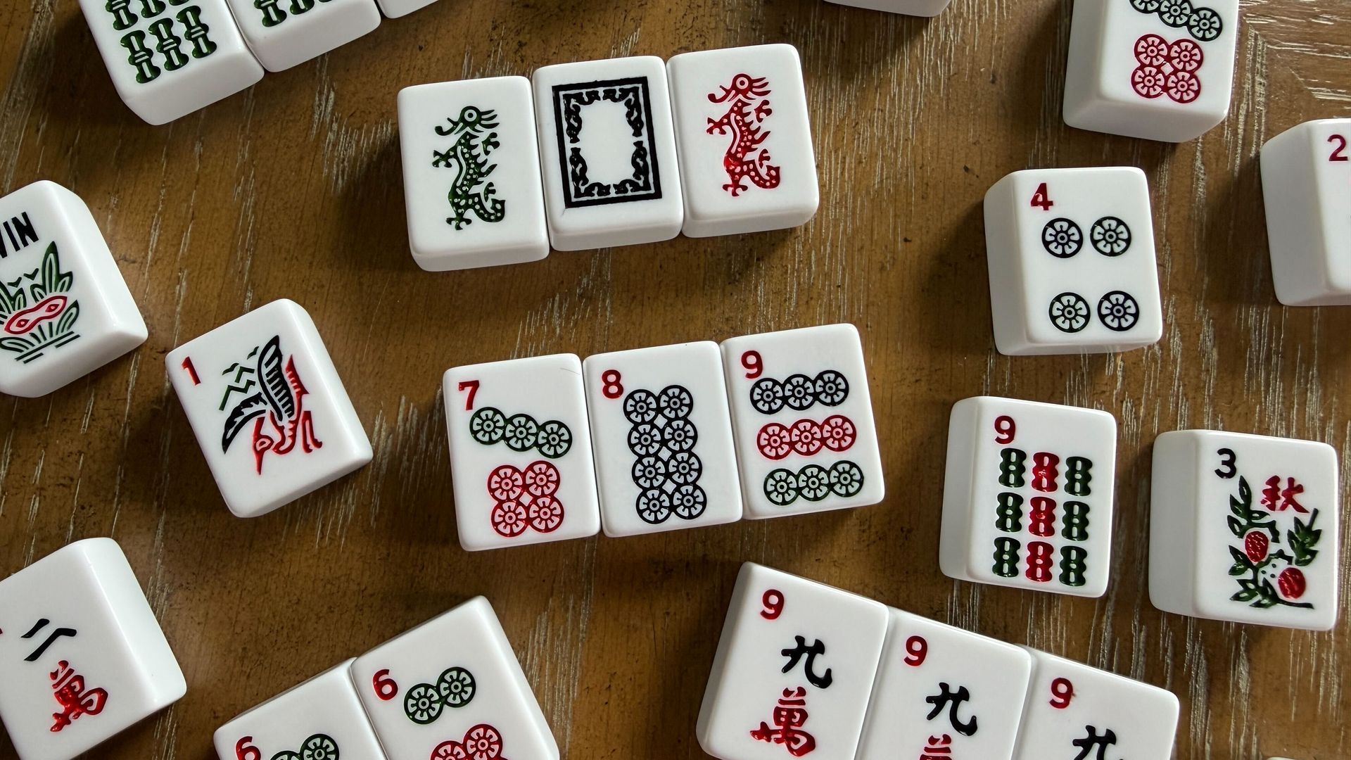Scattered white Mahjong tiles with colorful patterns including numbers, circles, dragons, flowers, and a bird on a brown wooden surface.