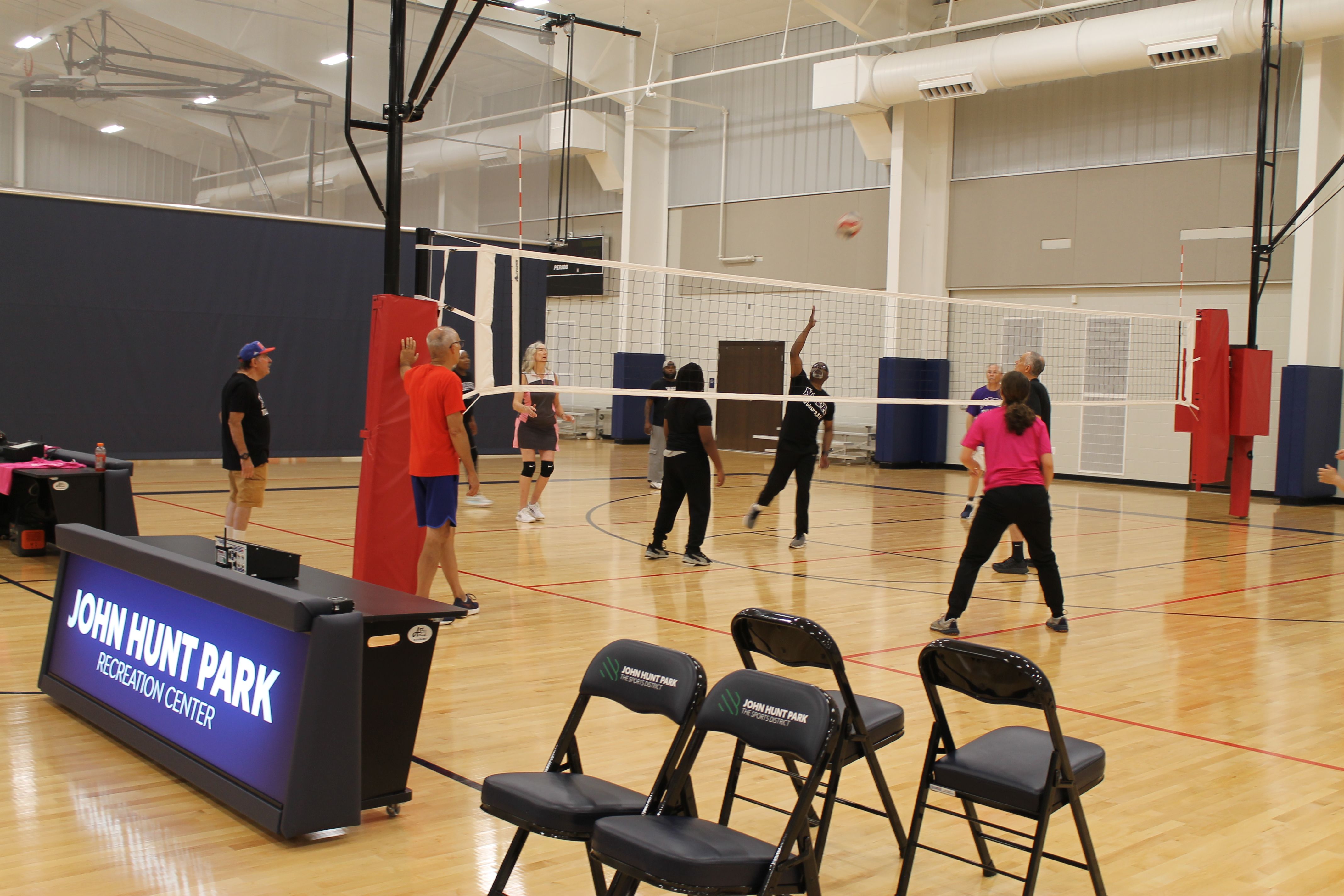Indoor gym volleyball game in progress; players near a net, one with raised hand ready to spike. In the foreground, chairs and a banner reading John Hunt Park Recreation Center.