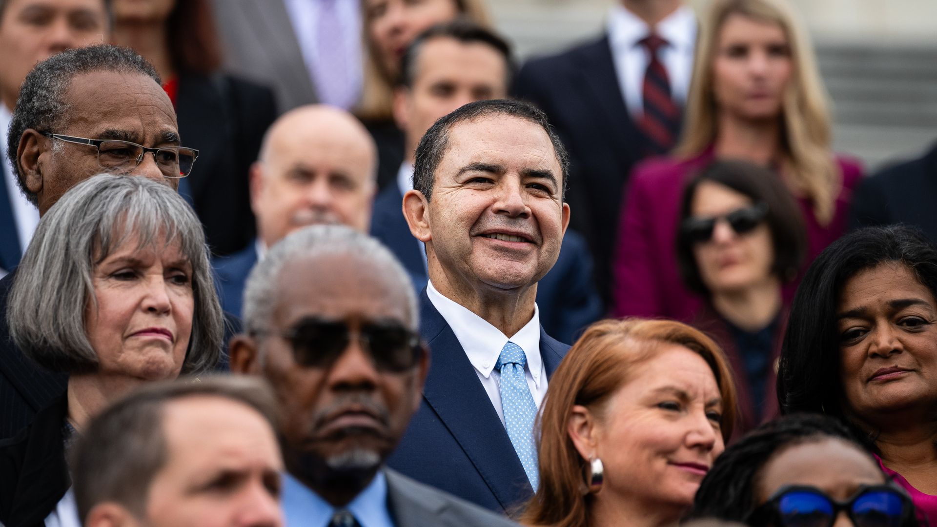 Group of people dressed in formal attire standing outdoors; man in center wears blue suit and light blue tie, smiling amid mostly serious expressions.