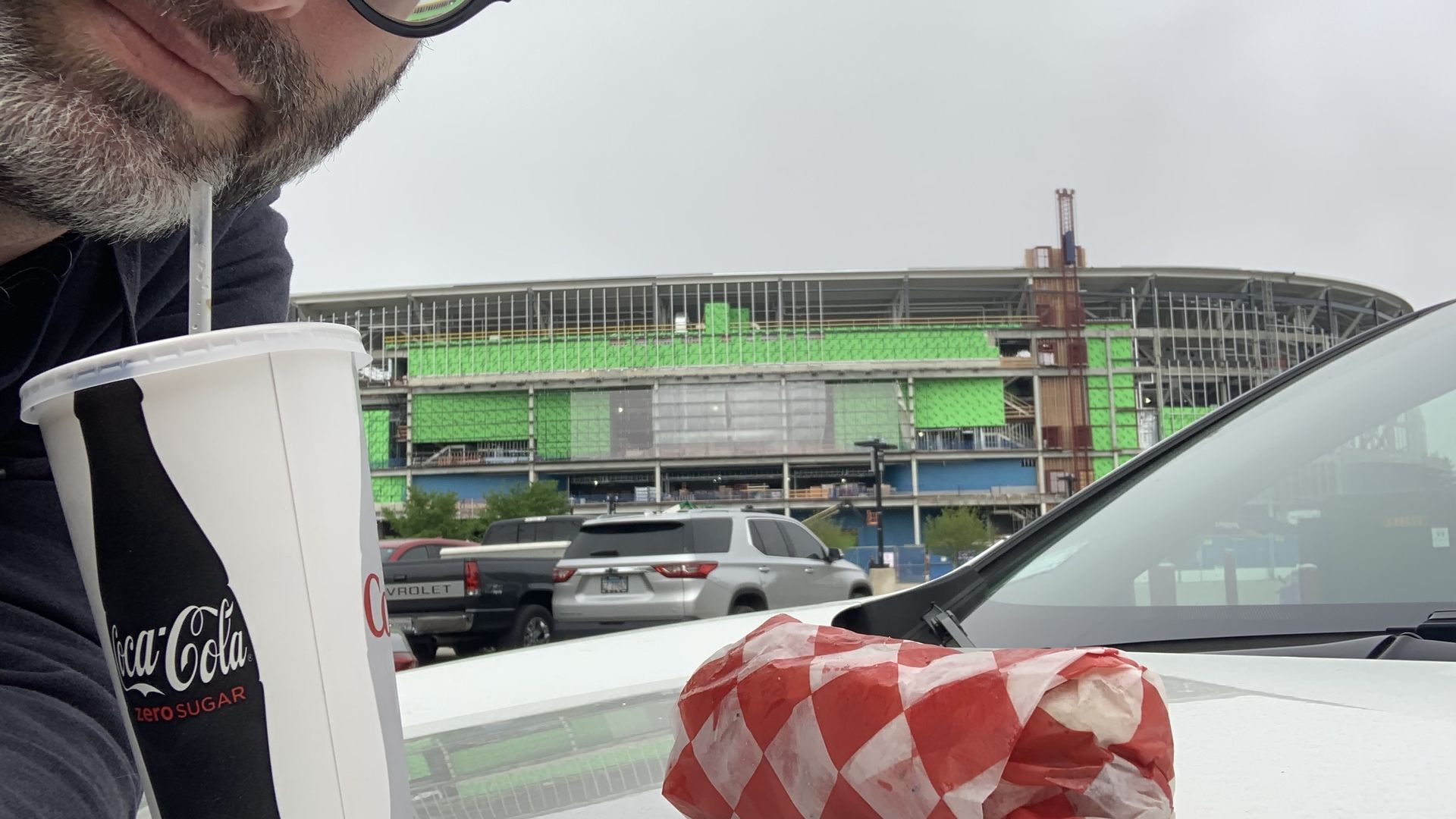 Bearded man in a black cap and glasses sips from a large Coca-Cola Zero Sugar cup with a straw; a construction site with green netting is in the background.