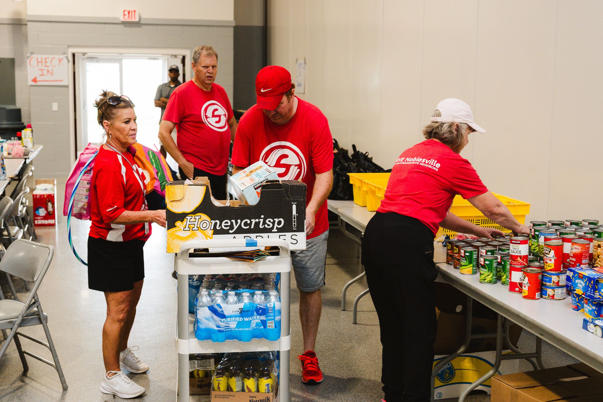 Volunteers in red shirts sort and bag food at a community drive. A Honeycrisp box on a cart, canned goods on the table, check-in sign and exit door in the background.