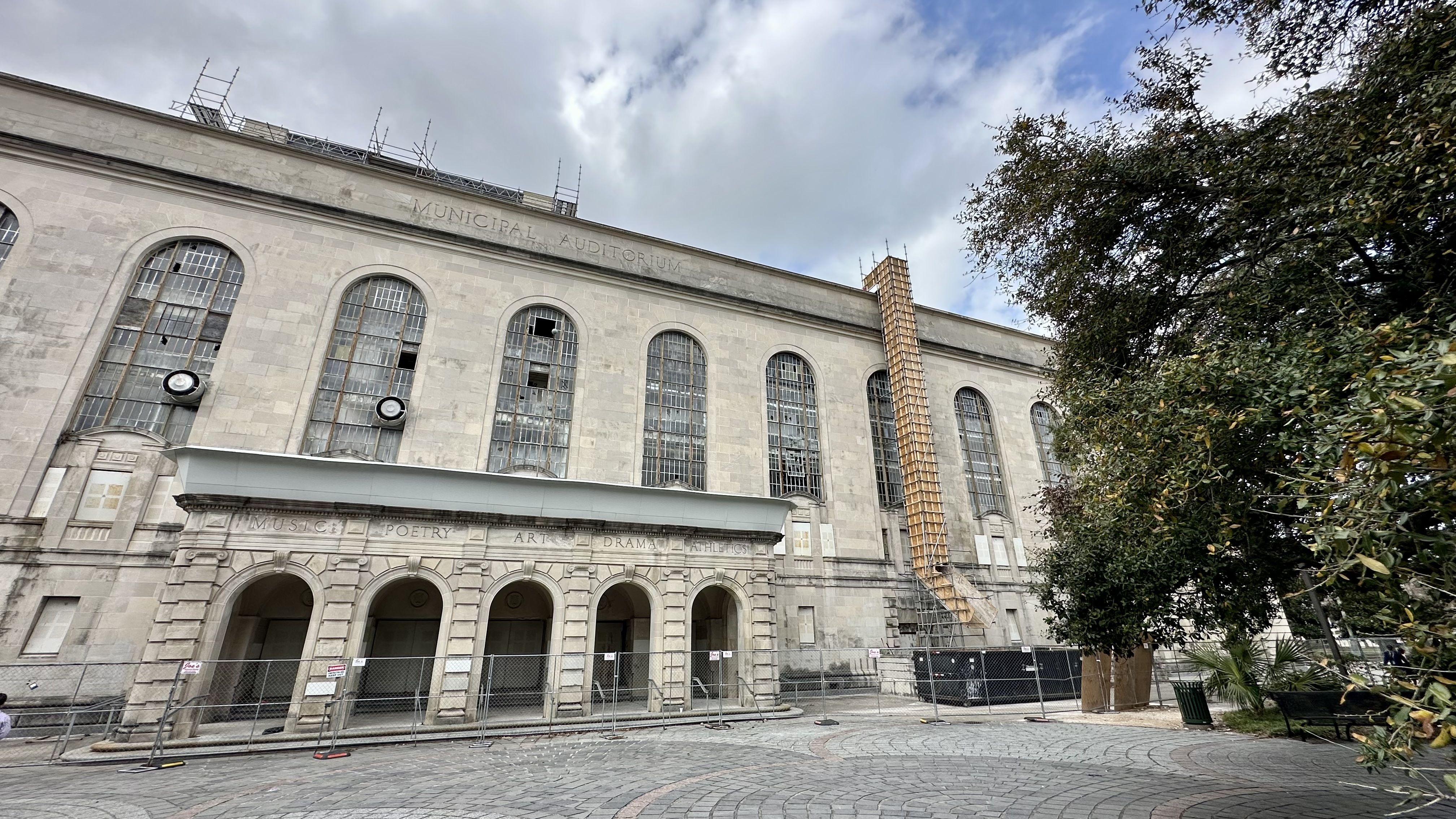 Old Municipal Auditorium building with tall arched windows, some broken, under a cloudy sky. Scaffolding and fencing indicate renovation. Trees and a curved stone path in foreground.