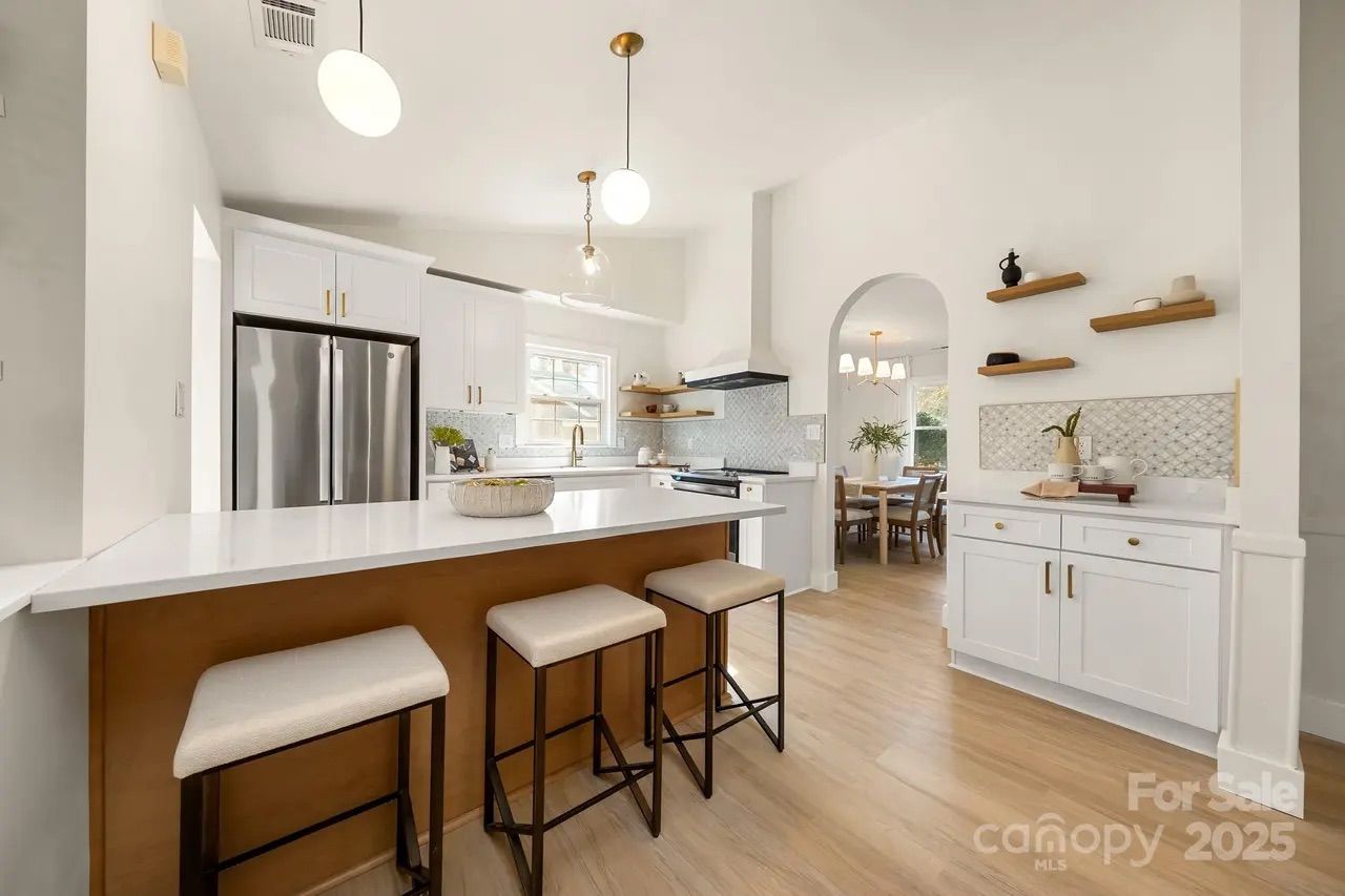 Bright kitchen with white cabinets, marble countertops, wooden bar with three beige cushioned stools, stainless steel fridge, pendant lights, and open archway to dining area with wooden table and chairs.
