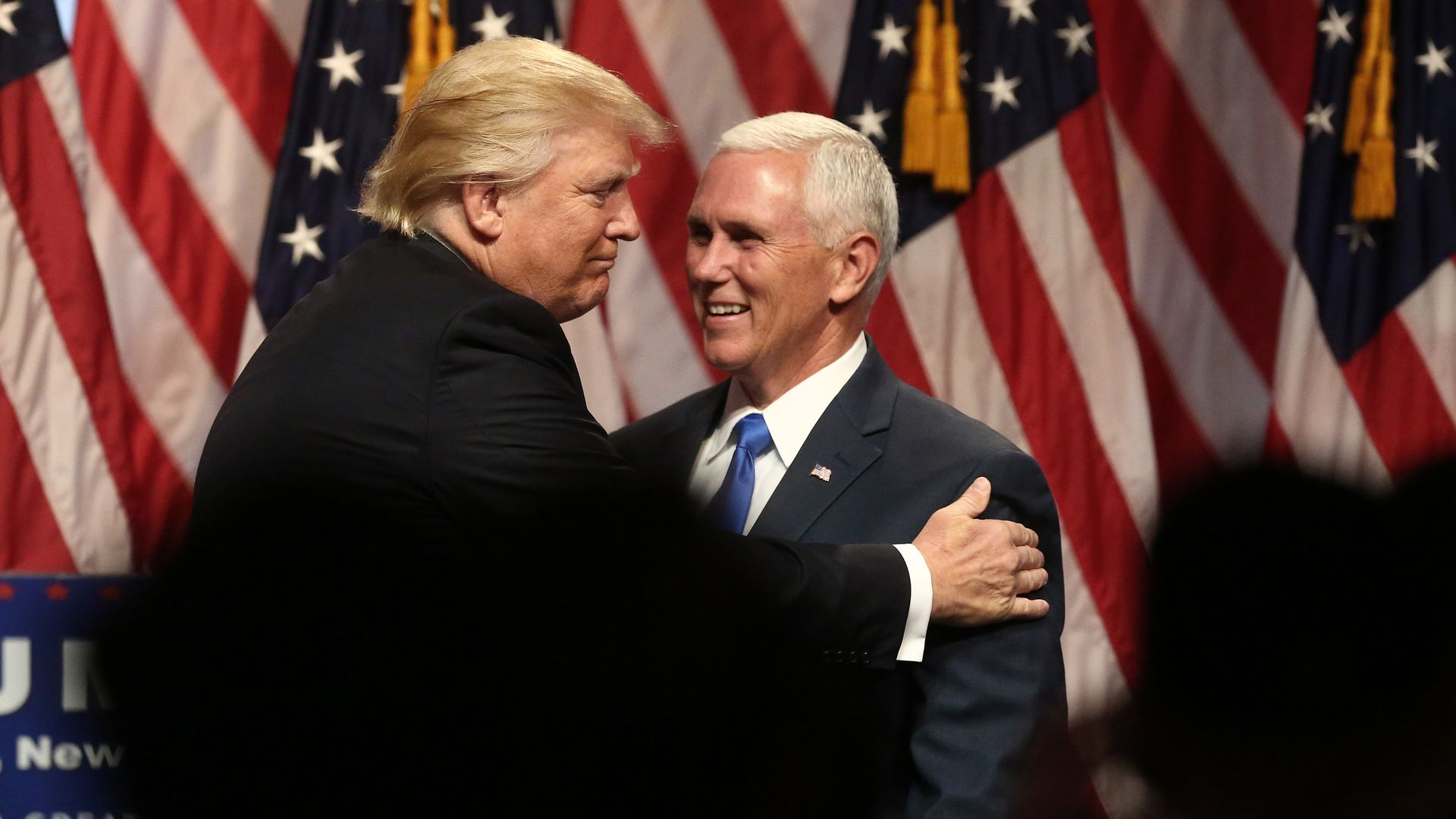 Two men in suits, one with blonde hair and one with white hair, embrace warmly in front of multiple American flags with red, white, and blue stripes and stars.