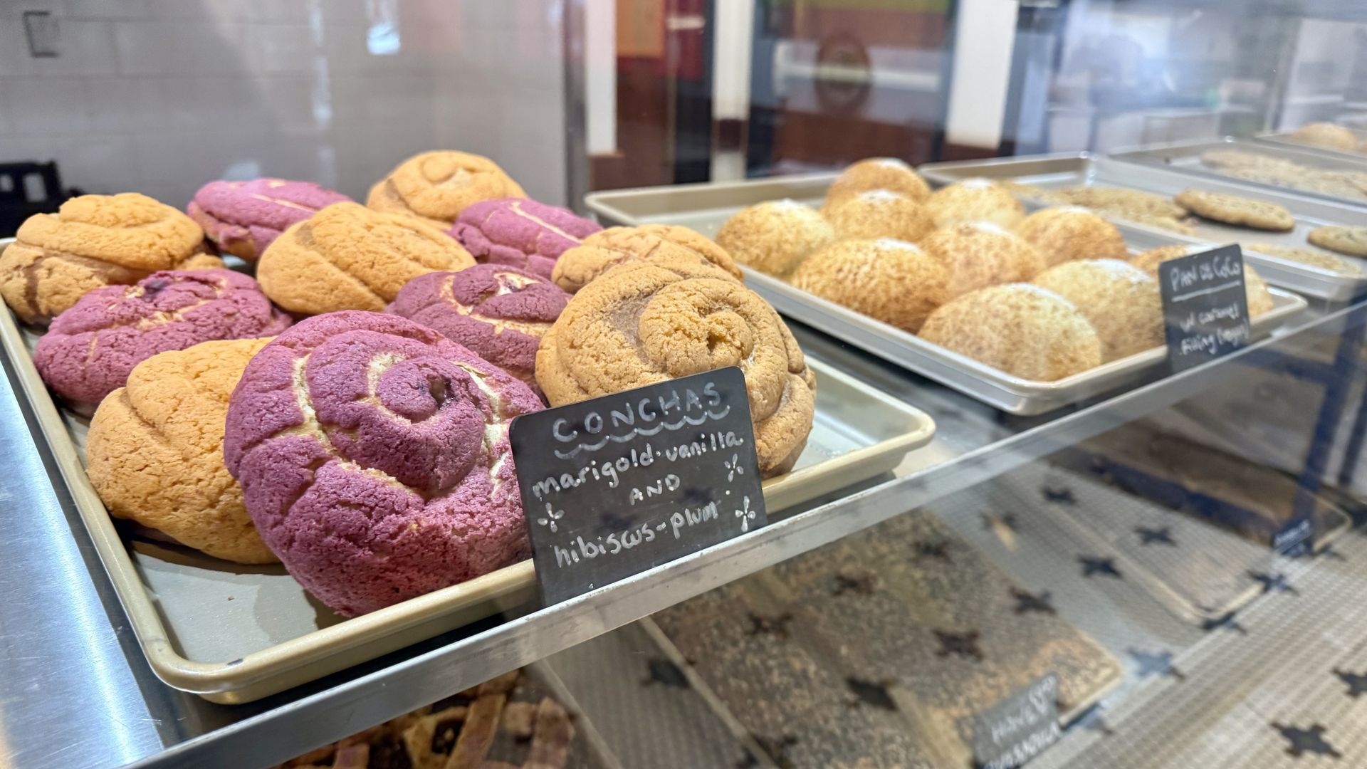Bakery display case showing trays of colorful conchas, a sweet bread, in orange and purple. Labels indicate flavors like marigold vanilla and hibiscus plum.