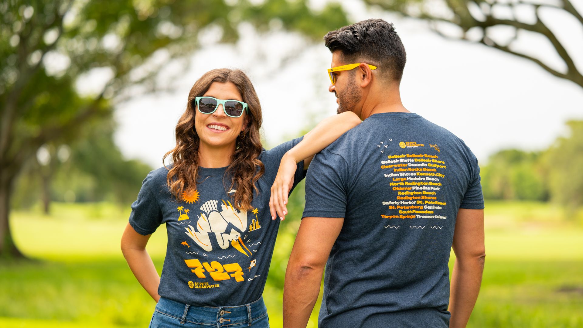 Two friends in a park wearing sunglasses and blue shirts that say "727 Day" in yellow with a drawing of a cream-colored pelican; woman with turquoise frames smiles at the camera, man with yellow frames faces away. Green trees blur in the background.