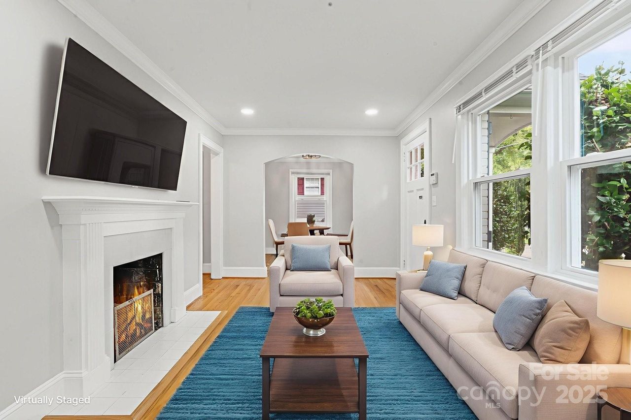Bright living room with beige sofa and armchair, blue rug, wooden coffee table with plant, white fireplace, large TV, and windows showing green foliage outside.