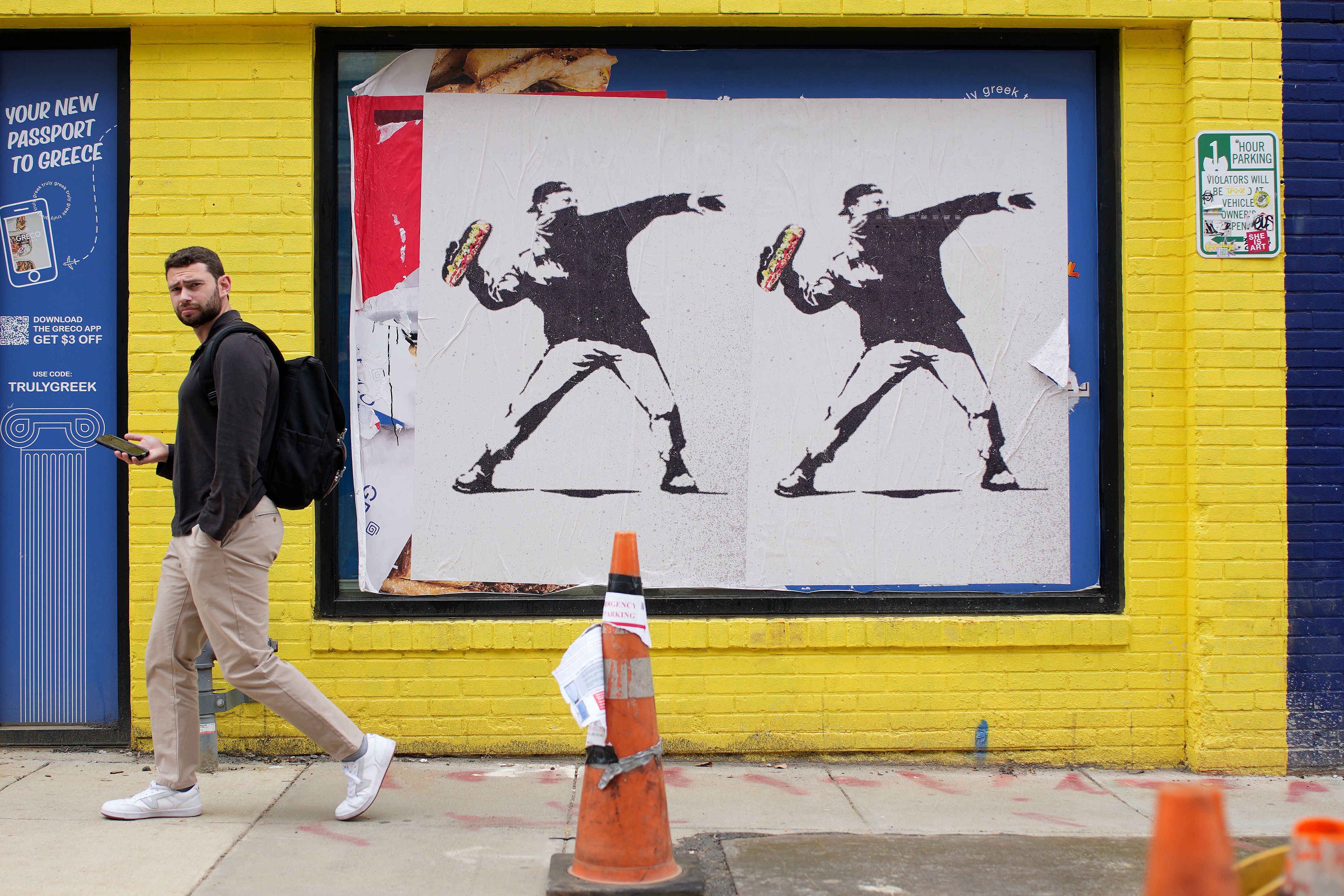 Man with backpack and phone walking past yellow brick wall with framed street art depicting two identical stencil figures throwing sandwiches.