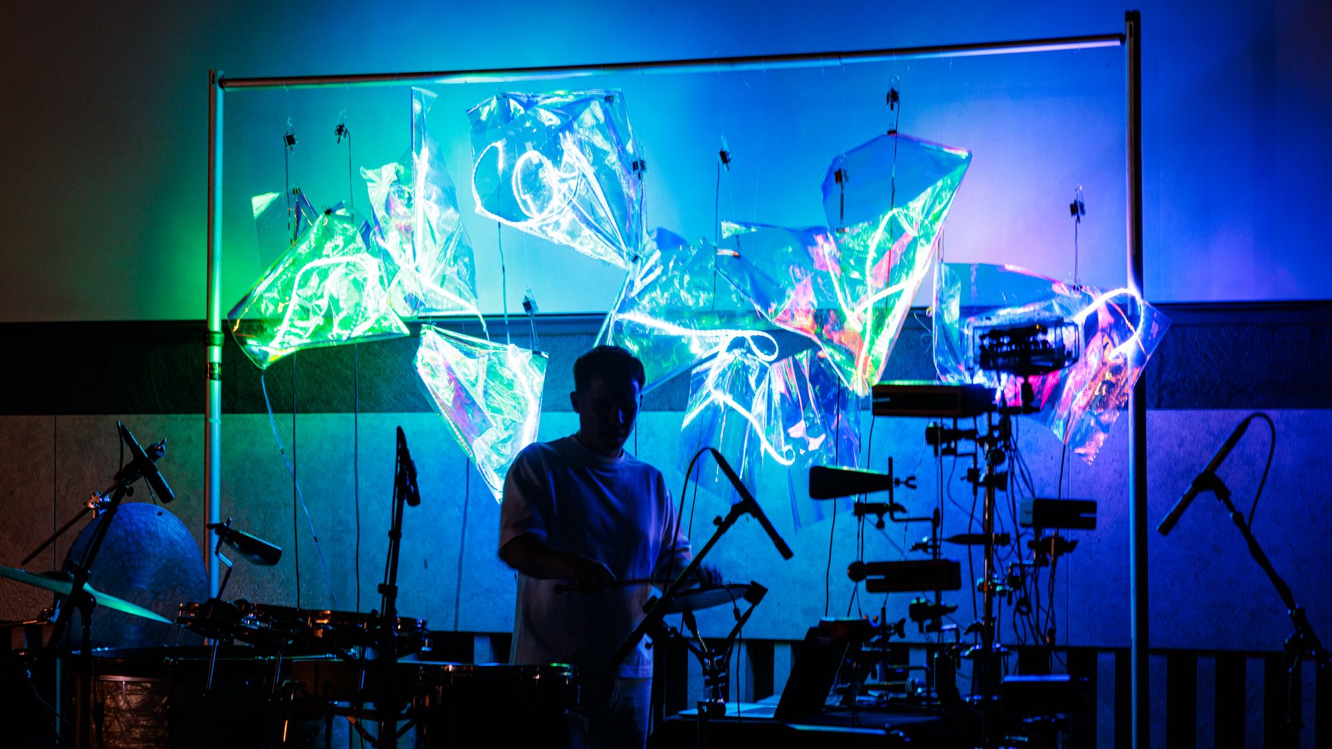 A man who is drumming is seen only in shadows while the backdrop is in colorful neon lights. 