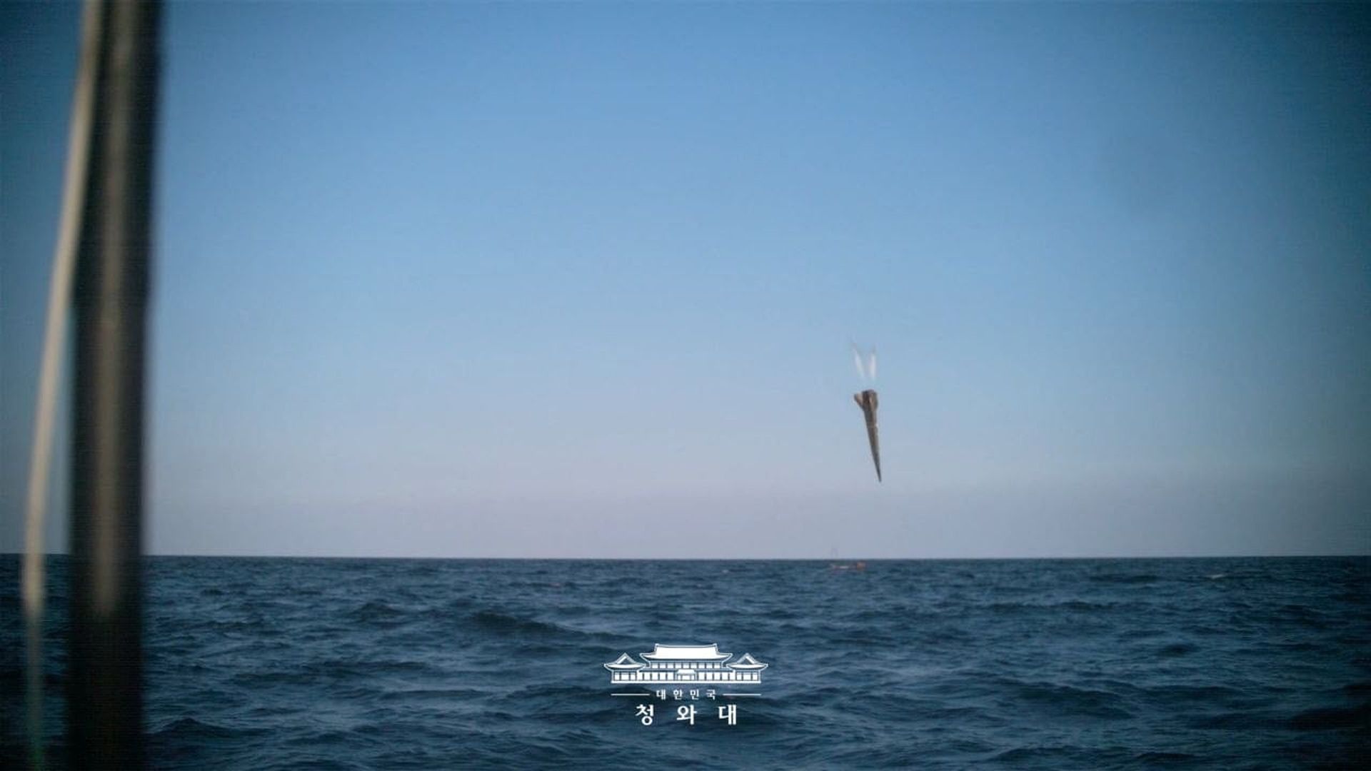 Blue ocean and pale sky meet at the horizon. A small, dark, kite-like shape hovers above the waves near center-right.