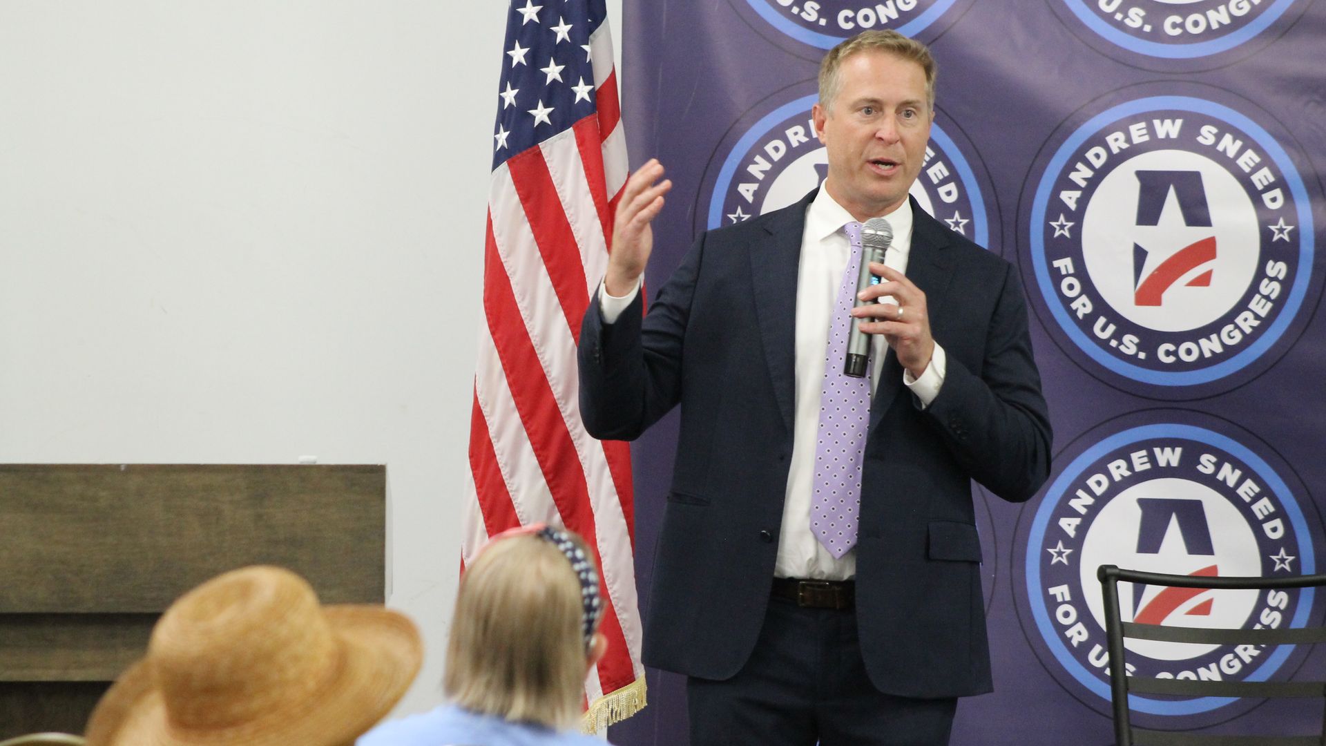 A man in a dark suit and lavender tie speaks into a microphone at a political event, gesturing with one hand; an American flag stands beside him and a purple backdrop bears circular campaign logos.
