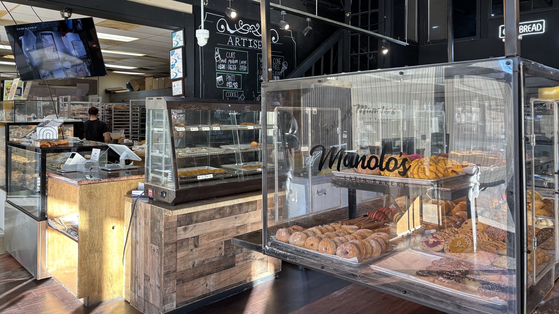 Interior of Manolo's Latin Bakery showing display cases with various pastries including donuts, cookies, and bread, wooden counters, and a chalkboard menu in the background.
