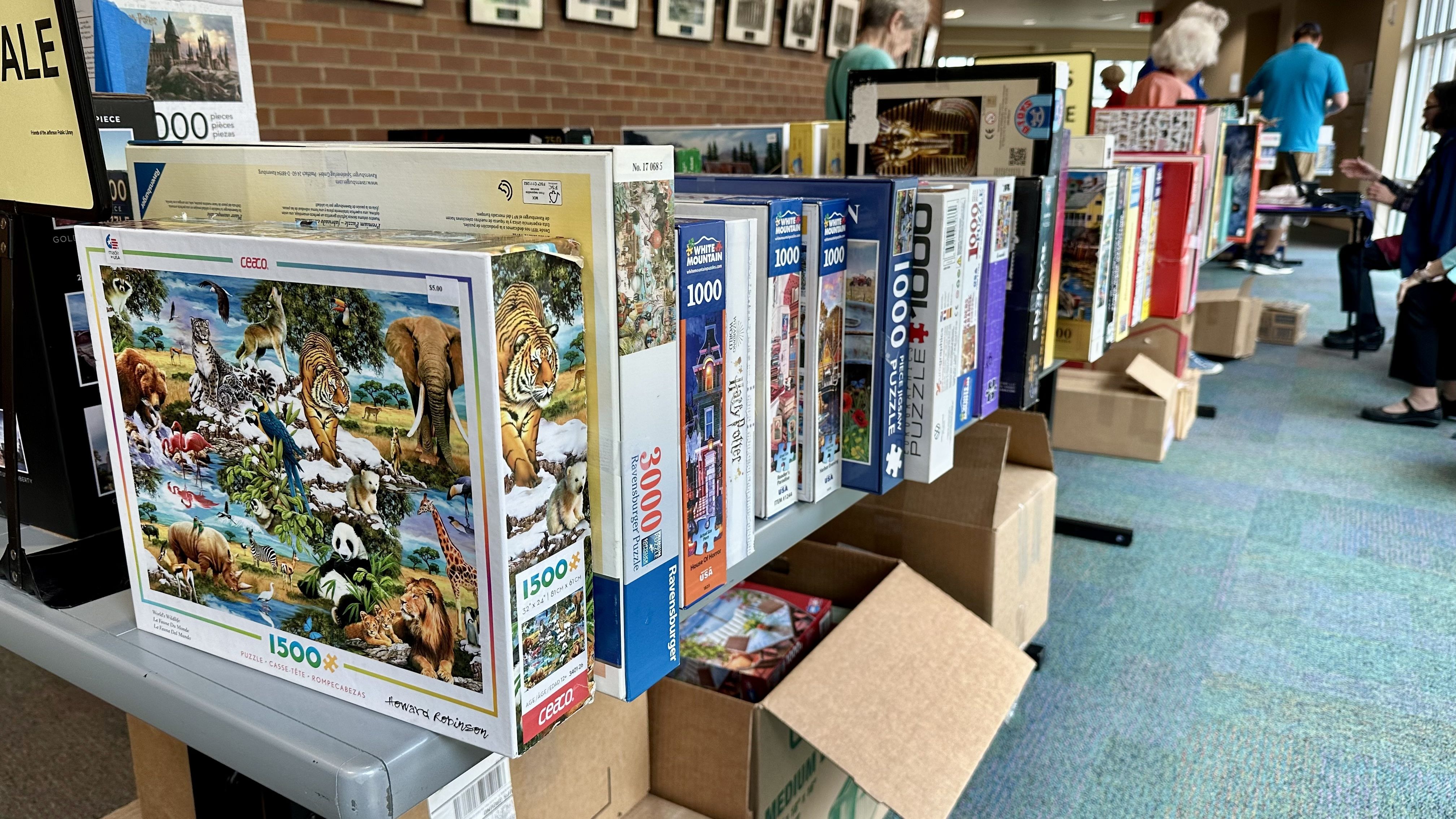 Colorful jigsaw puzzle boxes line a display table at a sale, set against a brick wall. Boxes show animal scenes with tigers and elephants; shoppers browse in the background.