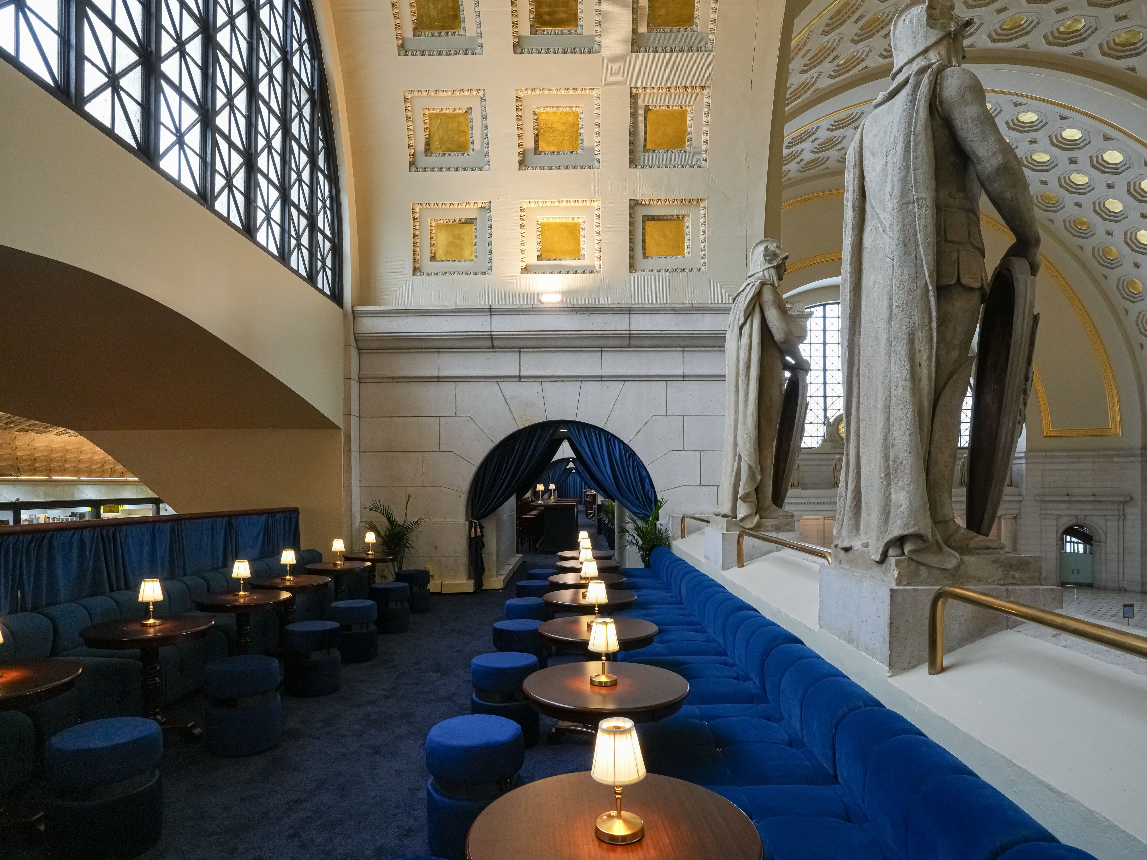 Interior of a grand museum hall: blue curved seating with small round lamps on tables, statues on a pedestal, gold-accented arched ceiling, and lattice windows along the left.