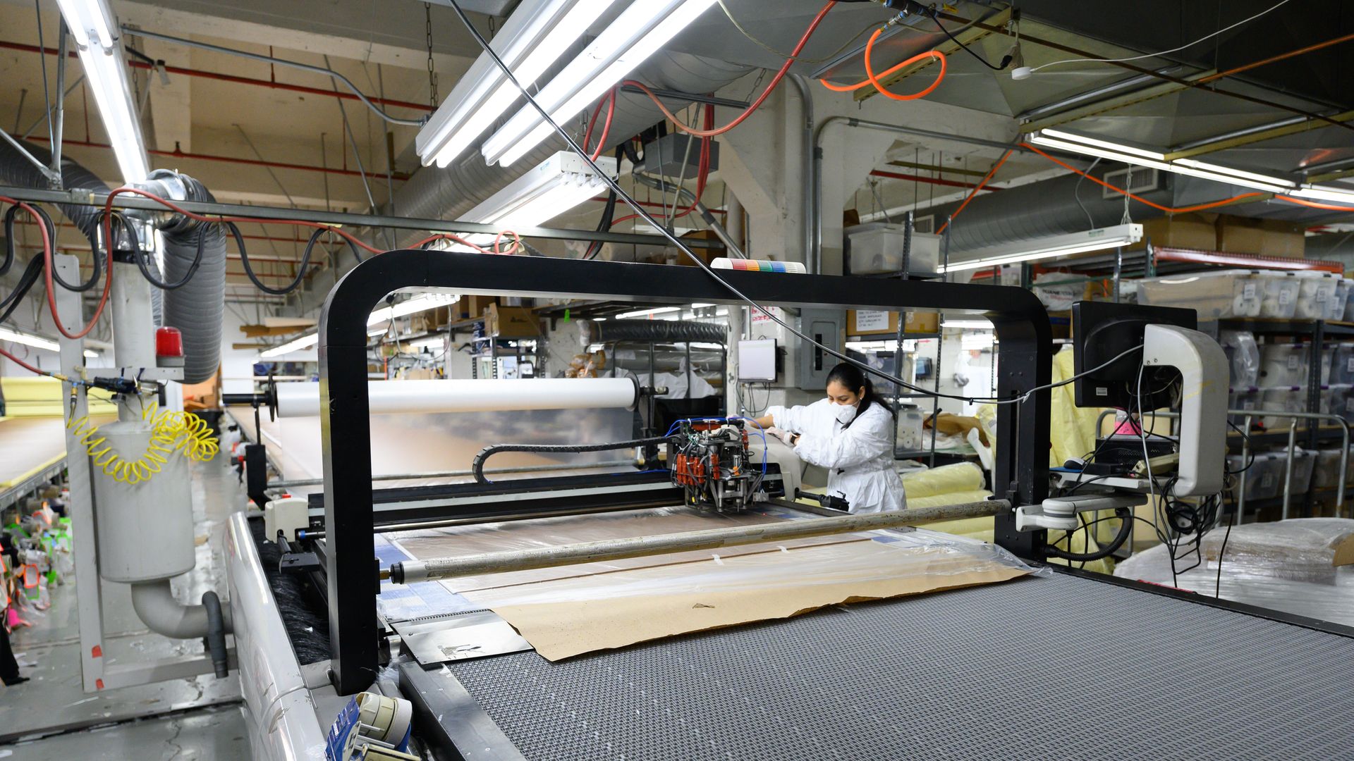 Masked worker standing in a factory at a large machine, cutting fabric 