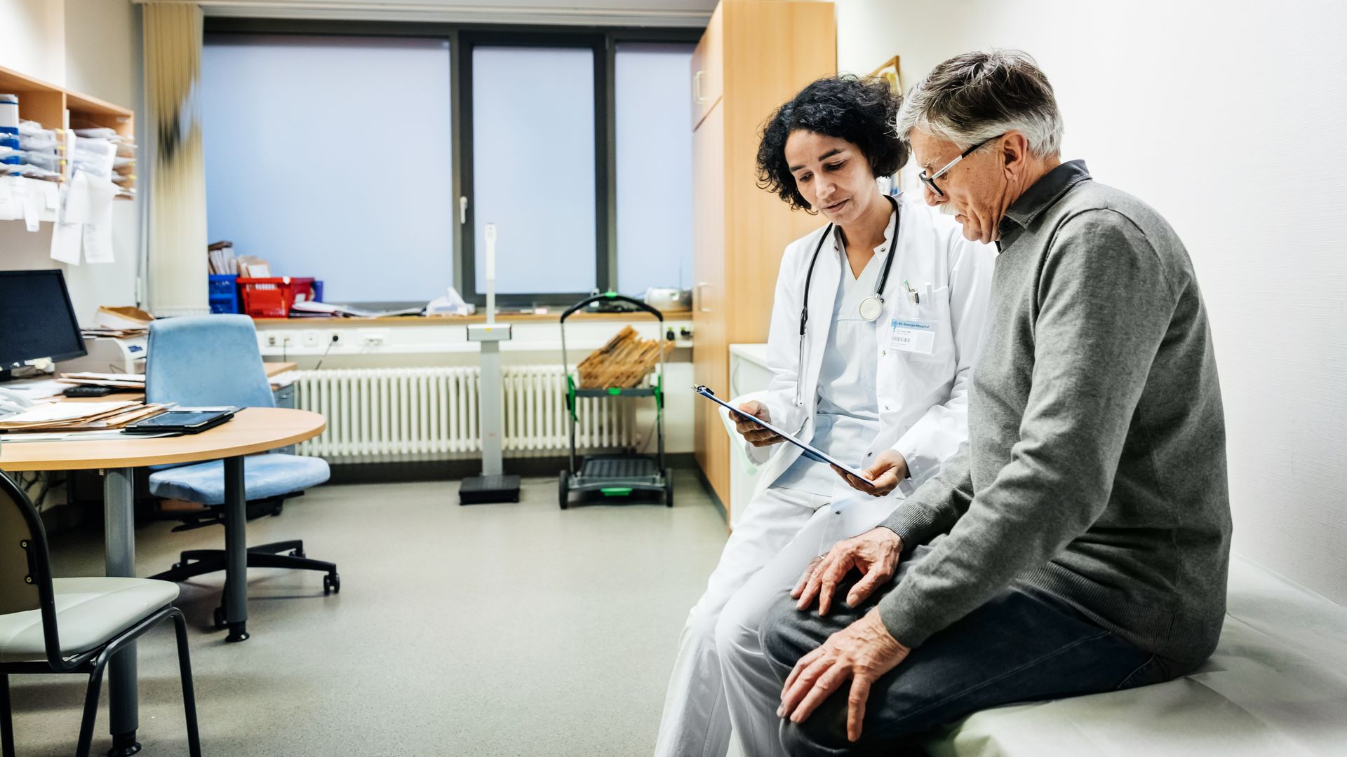 In this image, an elderly man in a gray sweater sits next to his doctor, a dark haired woman holding a clipboard.