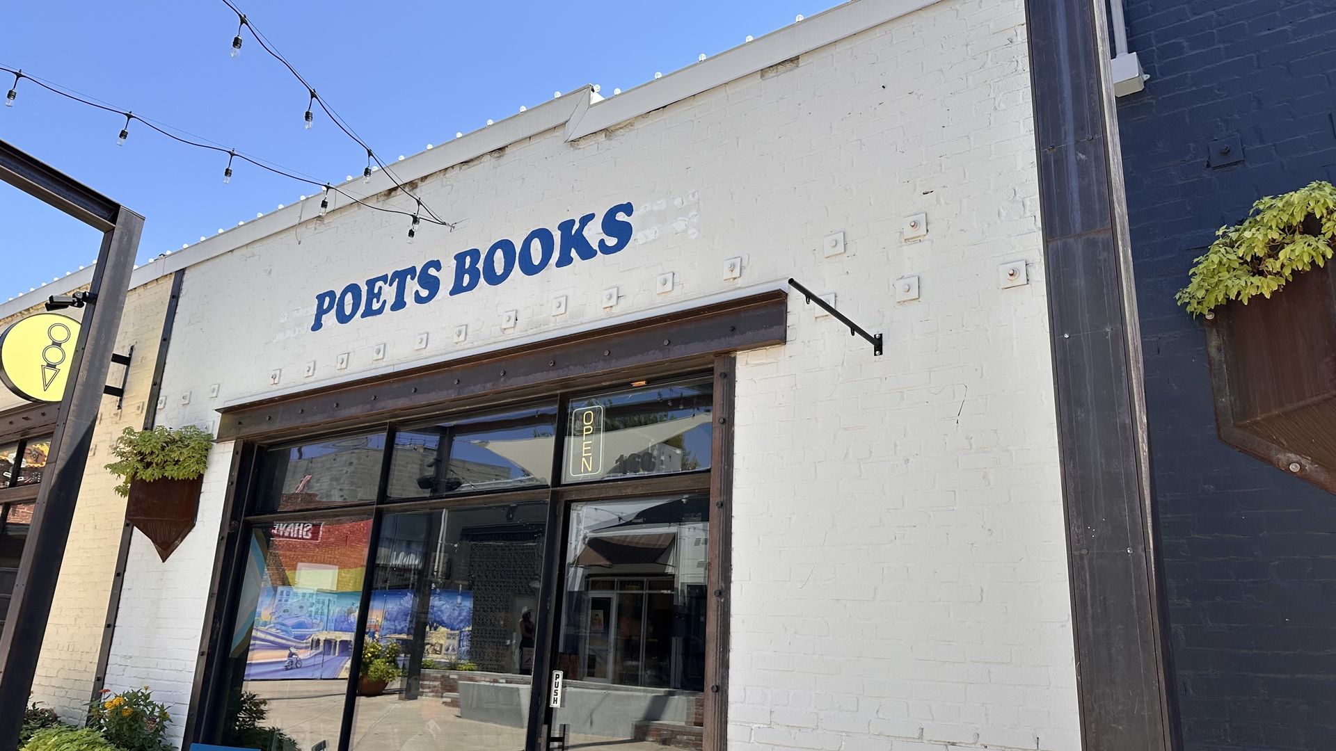 White brick building with blue text "POETS BOOKS" above large glass door. Door has a neon "OPEN" sign. String lights hang above entrance on a clear sunny day.