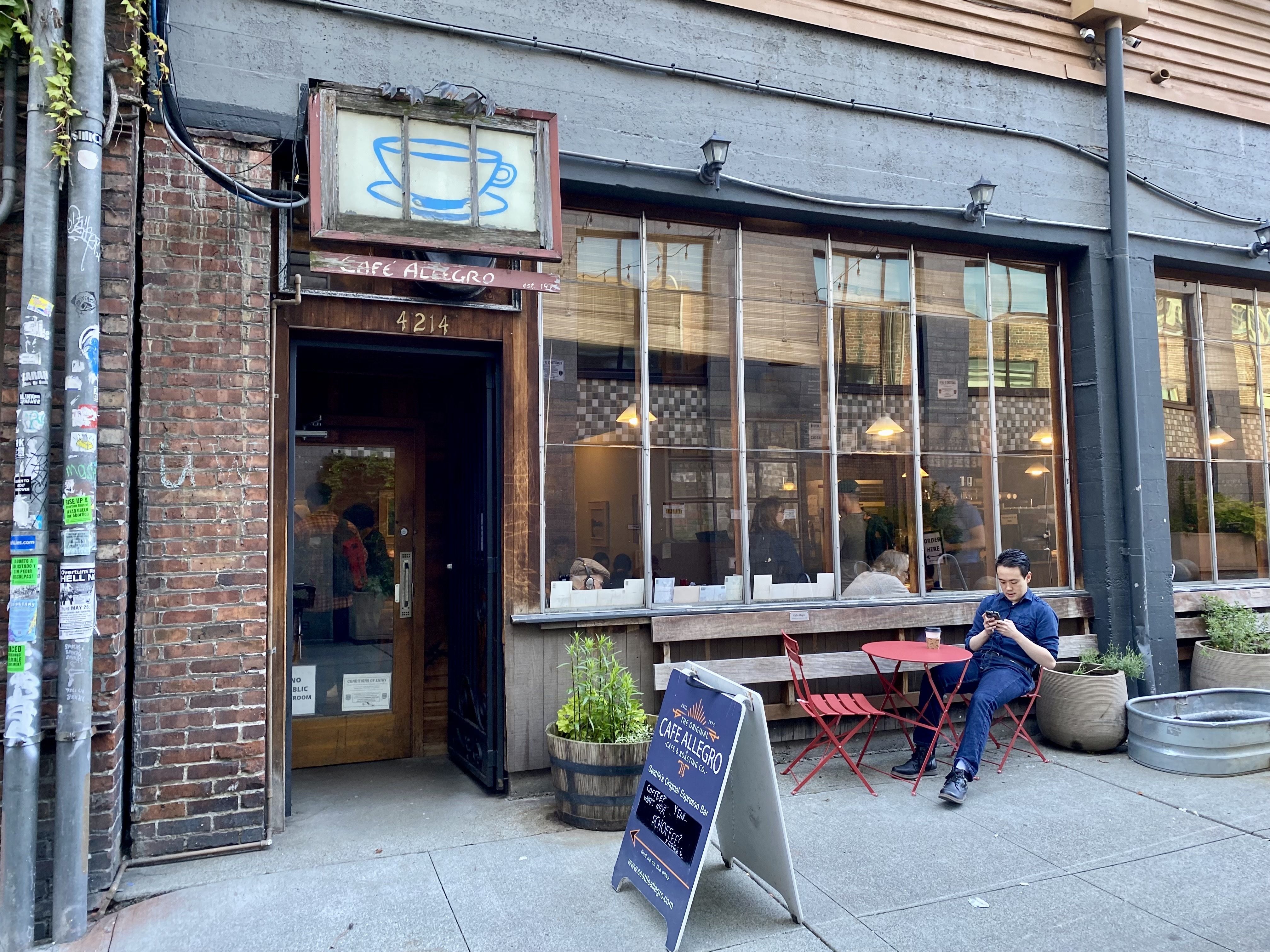 Entrance of Café Allegro with a blue coffee cup sign, red chairs and table outside, a man sitting using his phone, and people visible inside through large windows.