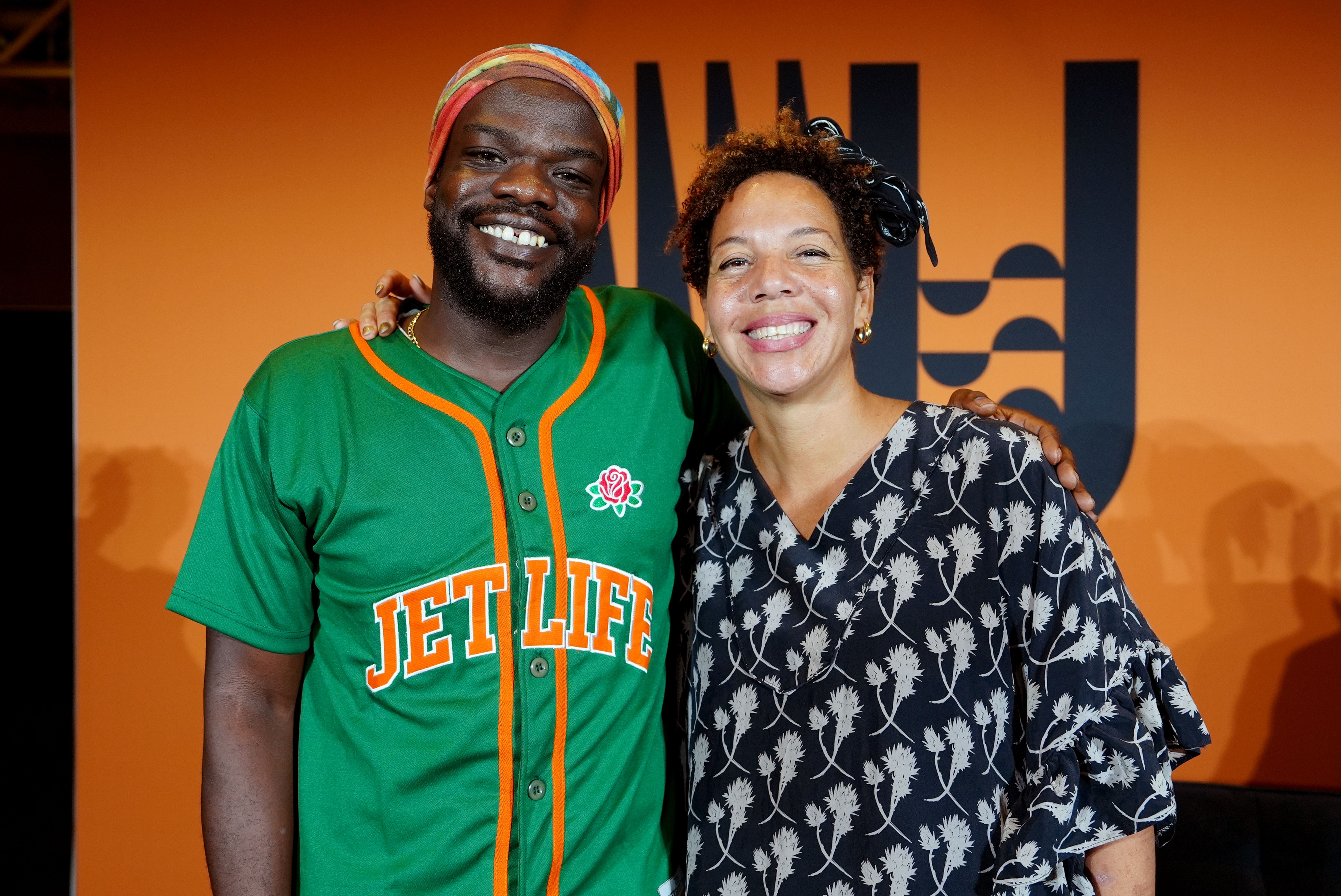 Smiling man in green "JET LIFE" shirt and colorful headwrap stands with smiling woman in blue floral dress with headband, orange background with partial black text behind them.