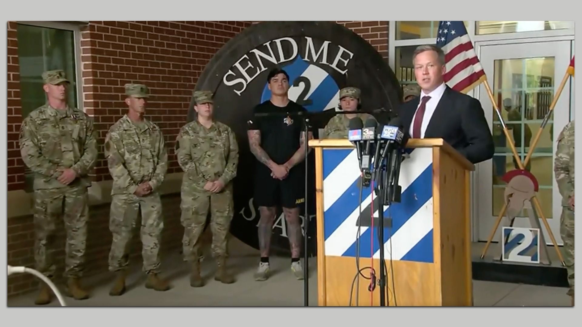 Military personnel stand beside a man in a suit speaking at a podium with microphones. Behind them is a large circular emblem with "SEND ME" text and the number 2, and an American flag.