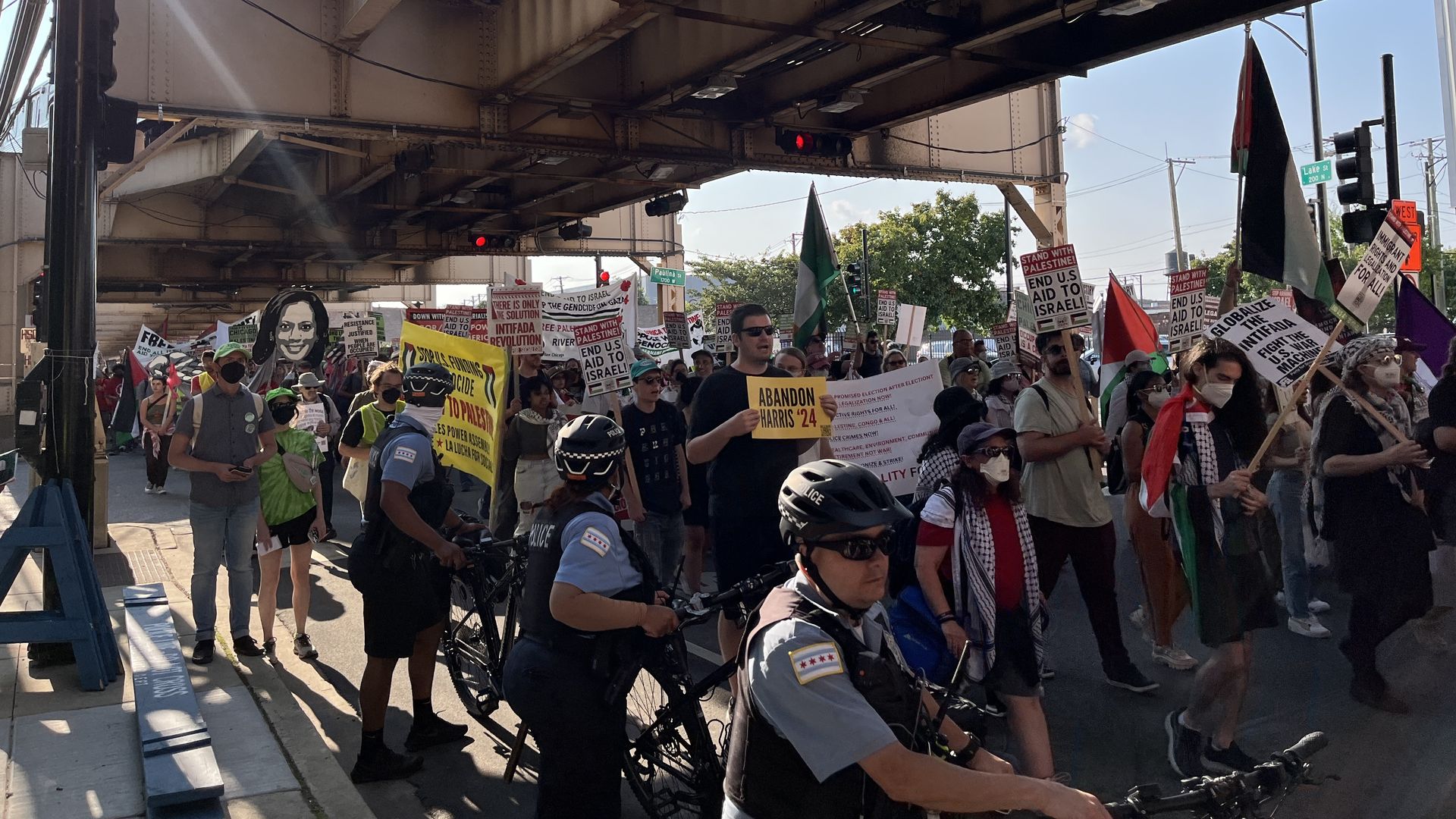 Protesters holding signs as bike cops line the street, all under elevated train tracks.