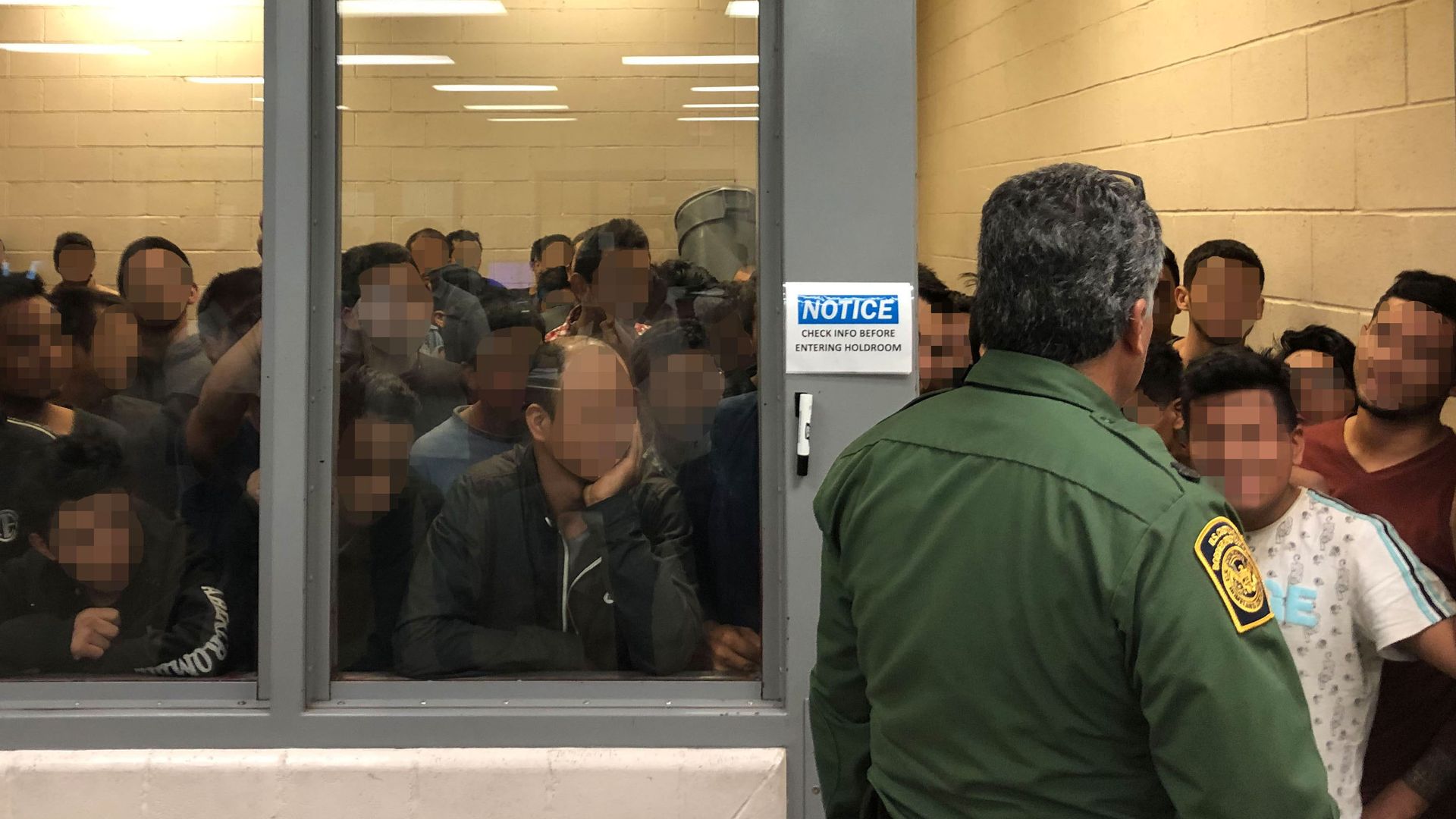 In this image, a CBP officer stands in front of a room of detained migrants