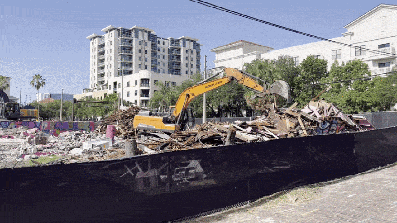Yellow excavator sits on a pile of construction rubble behind a black perimeter barrier; white modern apartment buildings and green trees rise in the background under a clear blue sky.