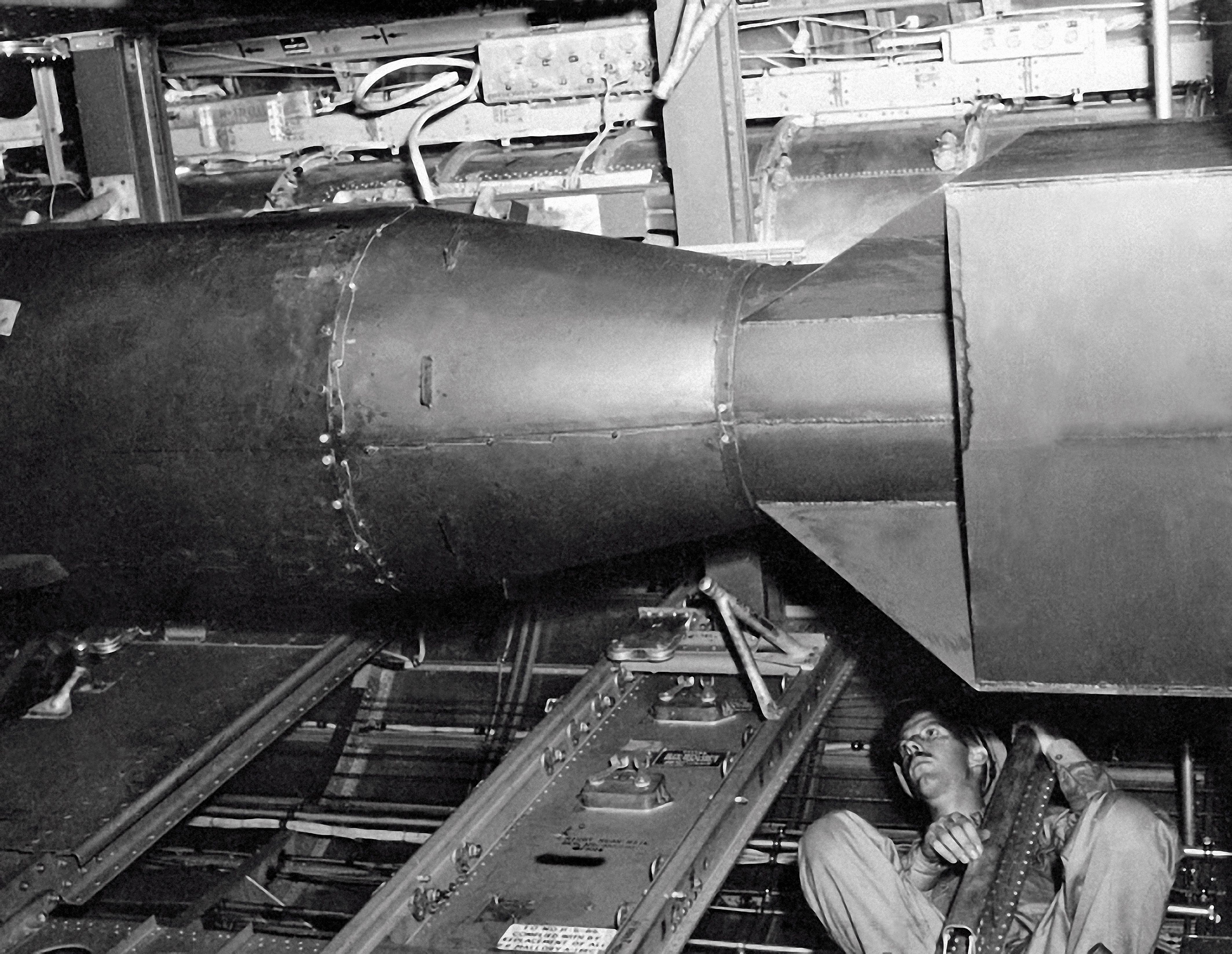 Black and white photo showing a man sitting beneath a large, cylindrical metal object known as the atomic bomb Little Boy with a conical end, surrounded by mechanical structures and cables inside an industrial or military setting.