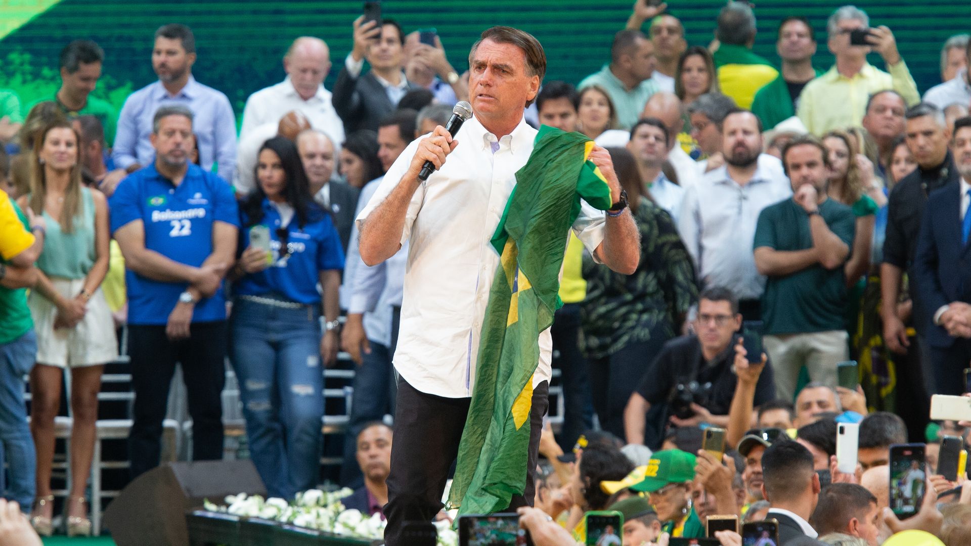 Brazil President Jair Bolsonaro holds up a Brazilian flag while speaking at a rally this weekend. He is surrounded by people 