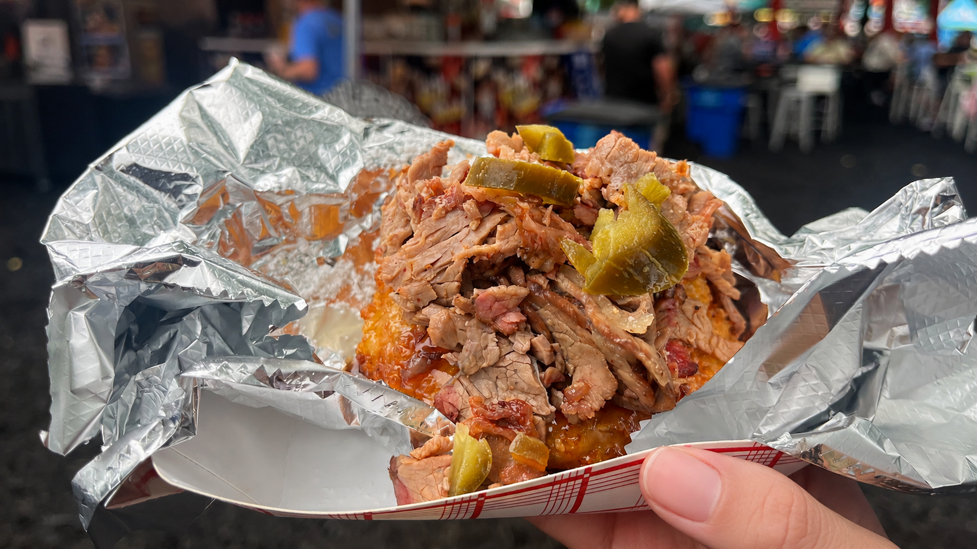 Close-up of a foil-wrapped sandwich piled with shredded barbecue meat and pickle slices, held in a hand with a busy outdoor market blurred in the background.