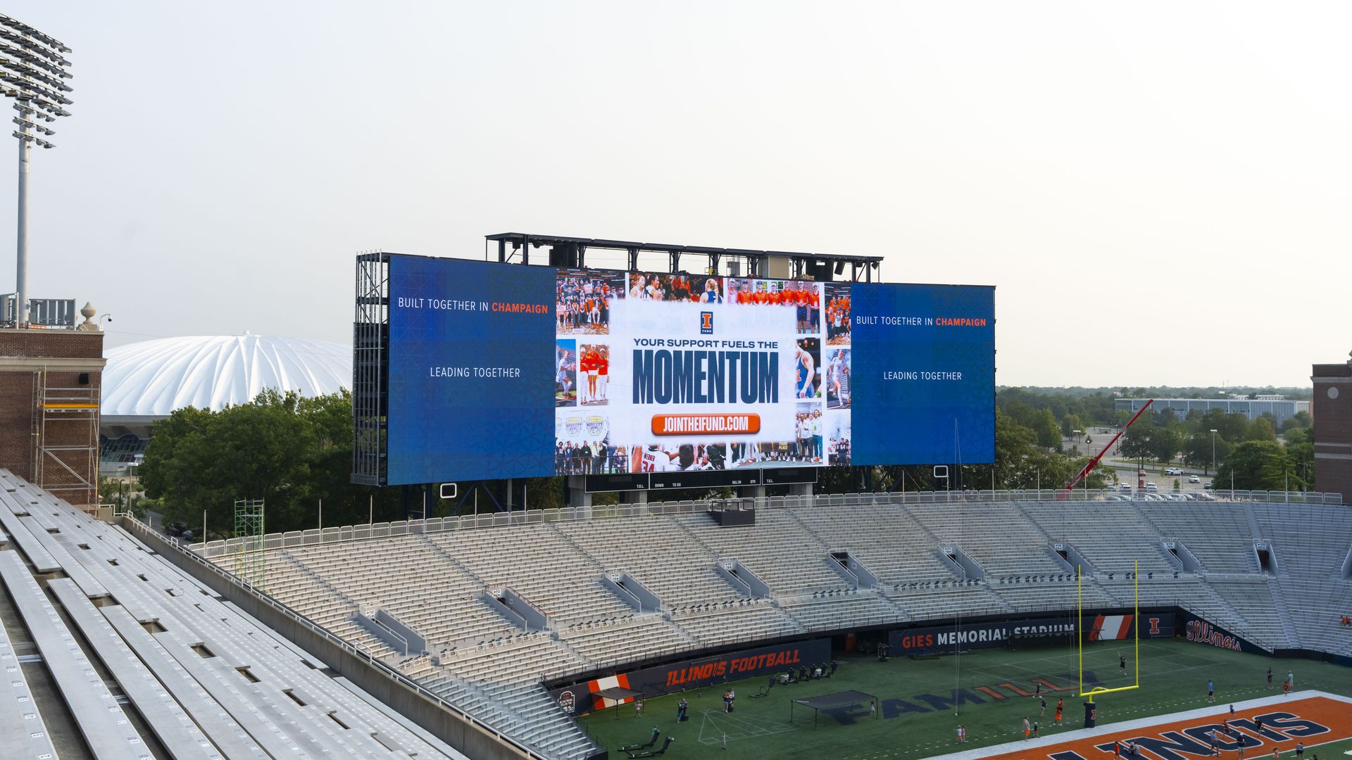 Wide stadium with a large blue scoreboard showing Momentum and Champaign, empty gray seating, a tall floodlight on the left, and a football field below under a pale sky.