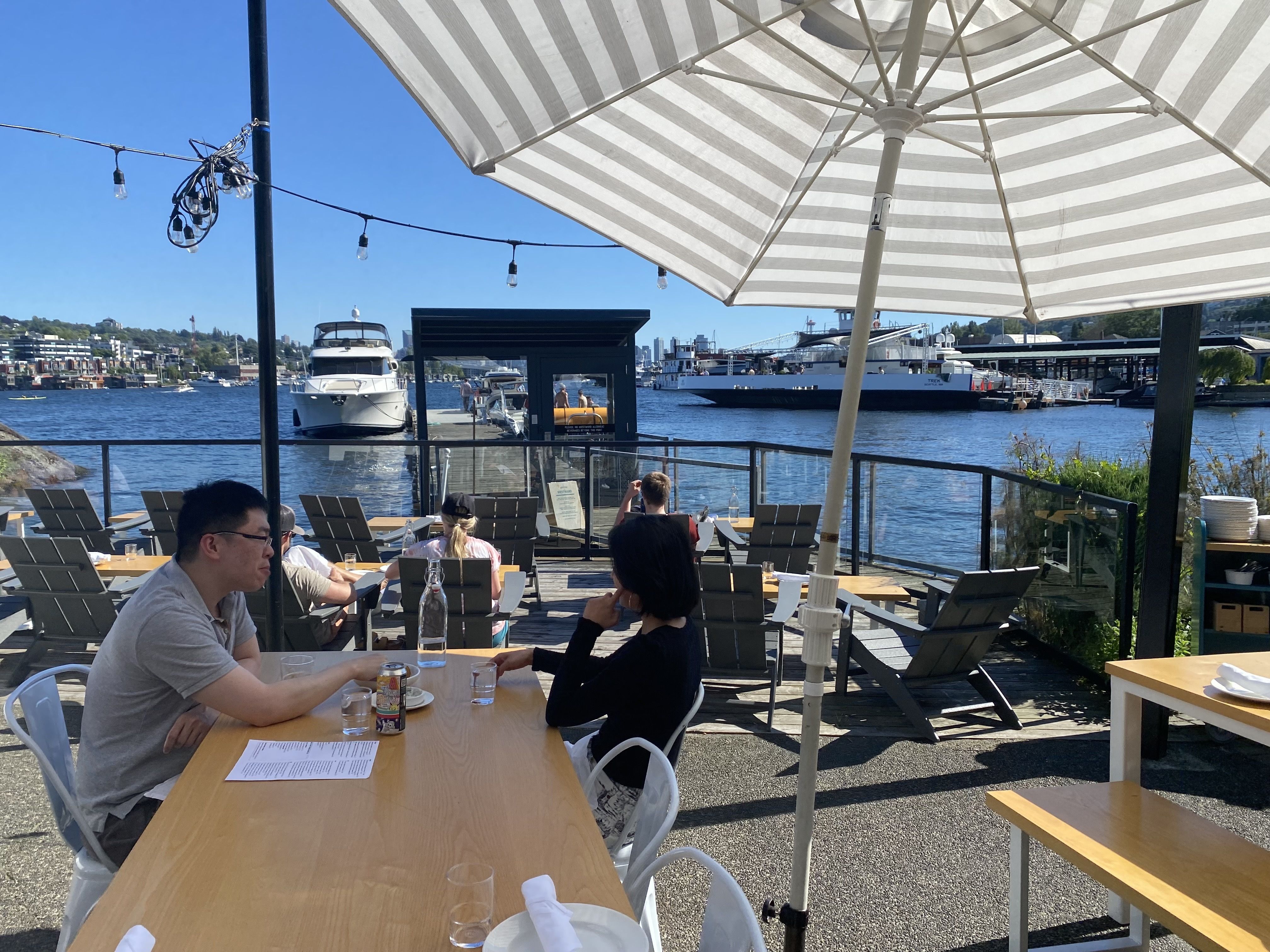 Outdoor waterfront dining area with wooden tables, gray chairs, and a large white and gray striped umbrella. People sit and relax near docks with boats and a large ferry on a sunny day.