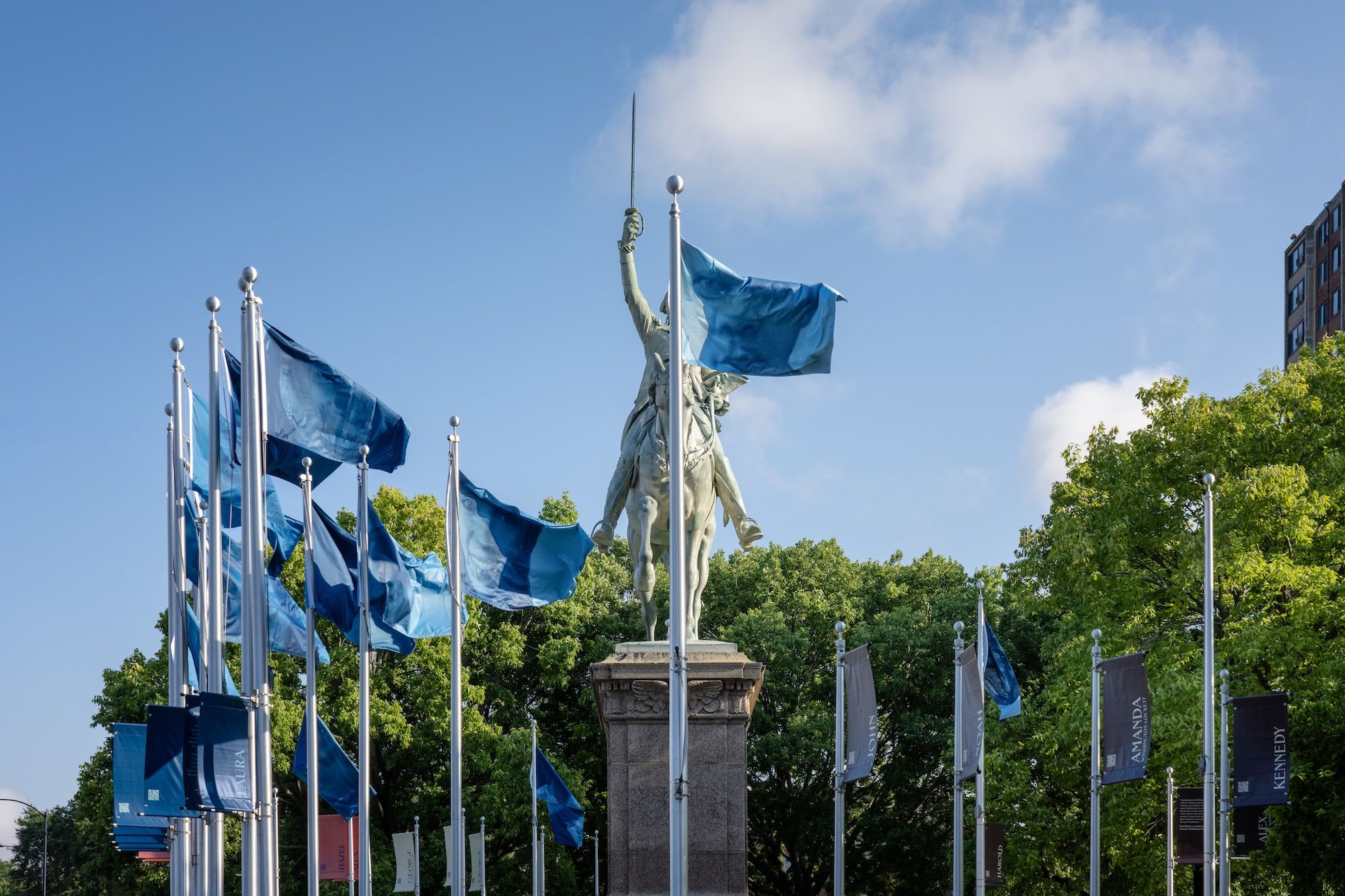 Statue raising a sword stands in a plaza, flanked by blue flagpoles; blue sky, green trees, and a building in the background.