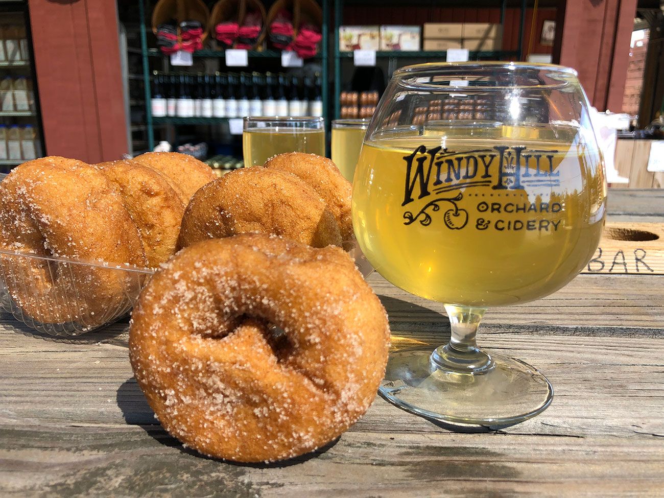 Sugar-coated mini donuts and a glass of light cider on a wooden table with a background of shelves holding bottles at Windy Hill Orchard & Cidery.