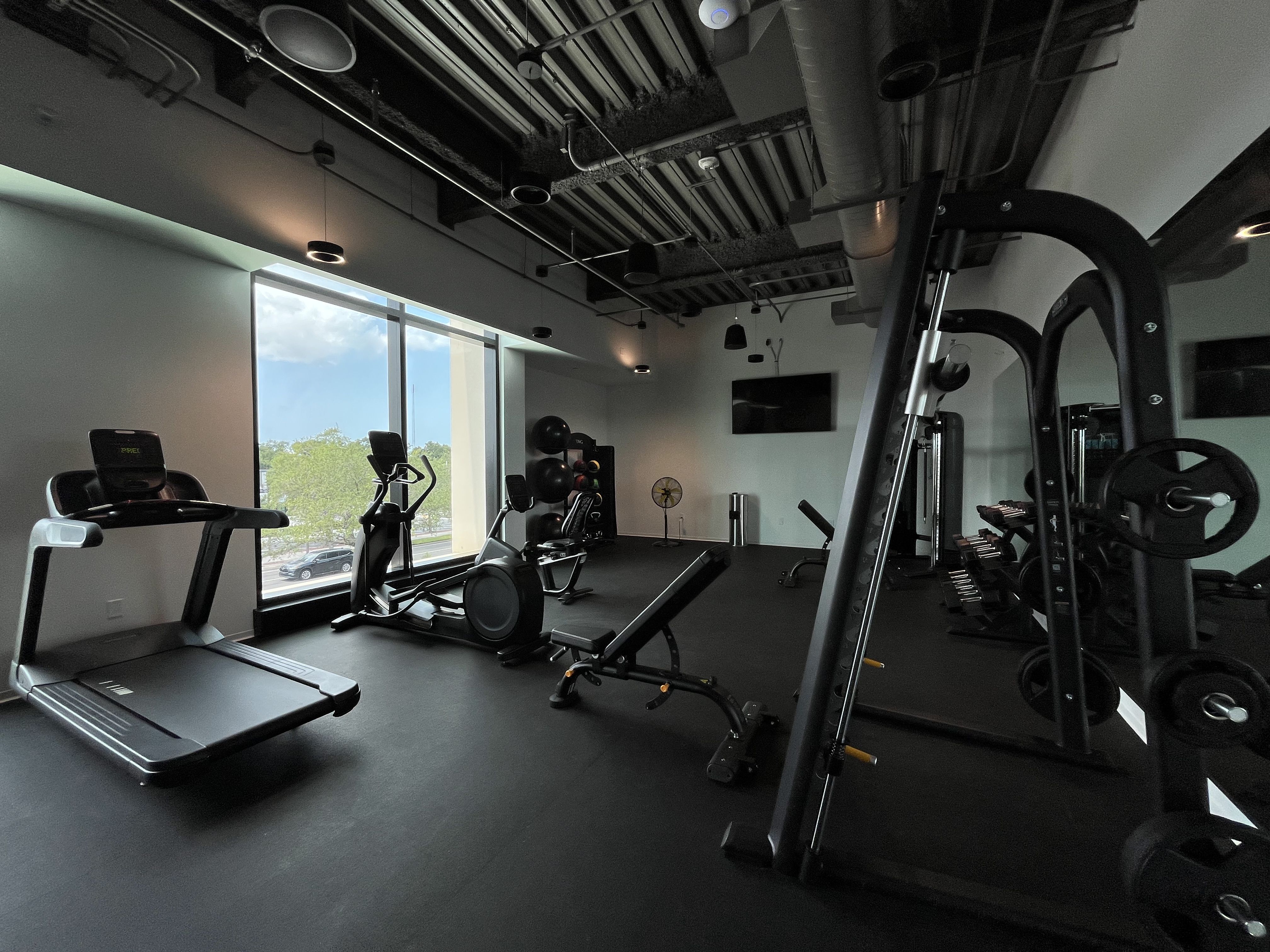 A modern gym interior with large left-side windows, a row of treadmills and ellipticals, exposed ceiling ducts, and weight machines in the background.
