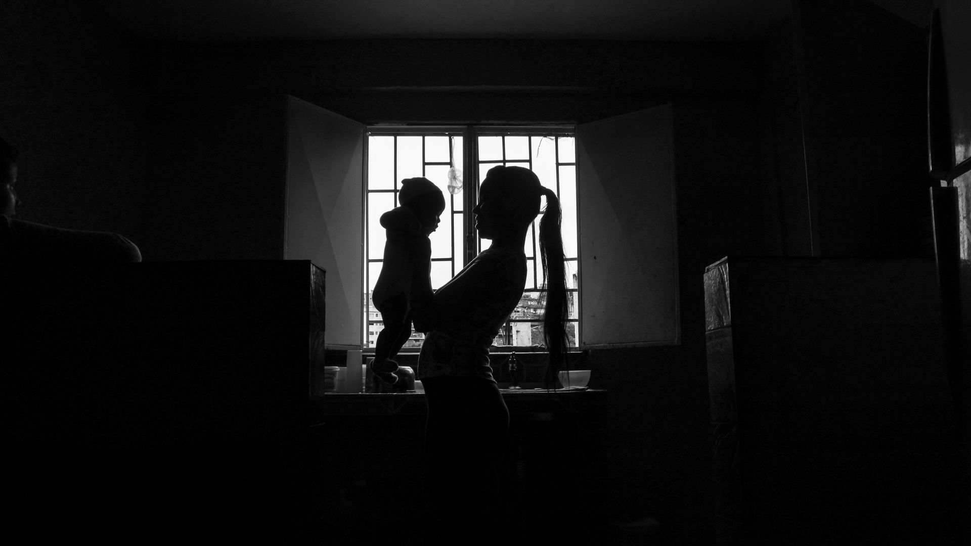 A mother holds her baby in front of a window in the shadows.