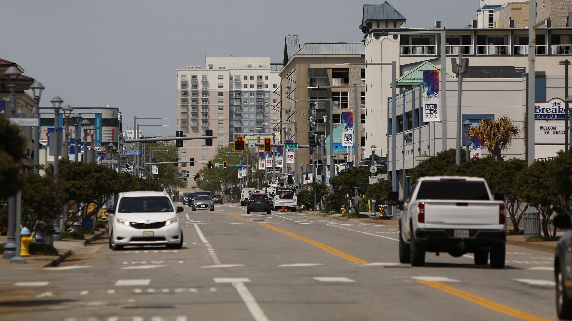 Busy city street lined with mid-rise buildings, traffic lights, and banners. White minivan left, white pickup right, cars in the distance, clear blue sky overhead.