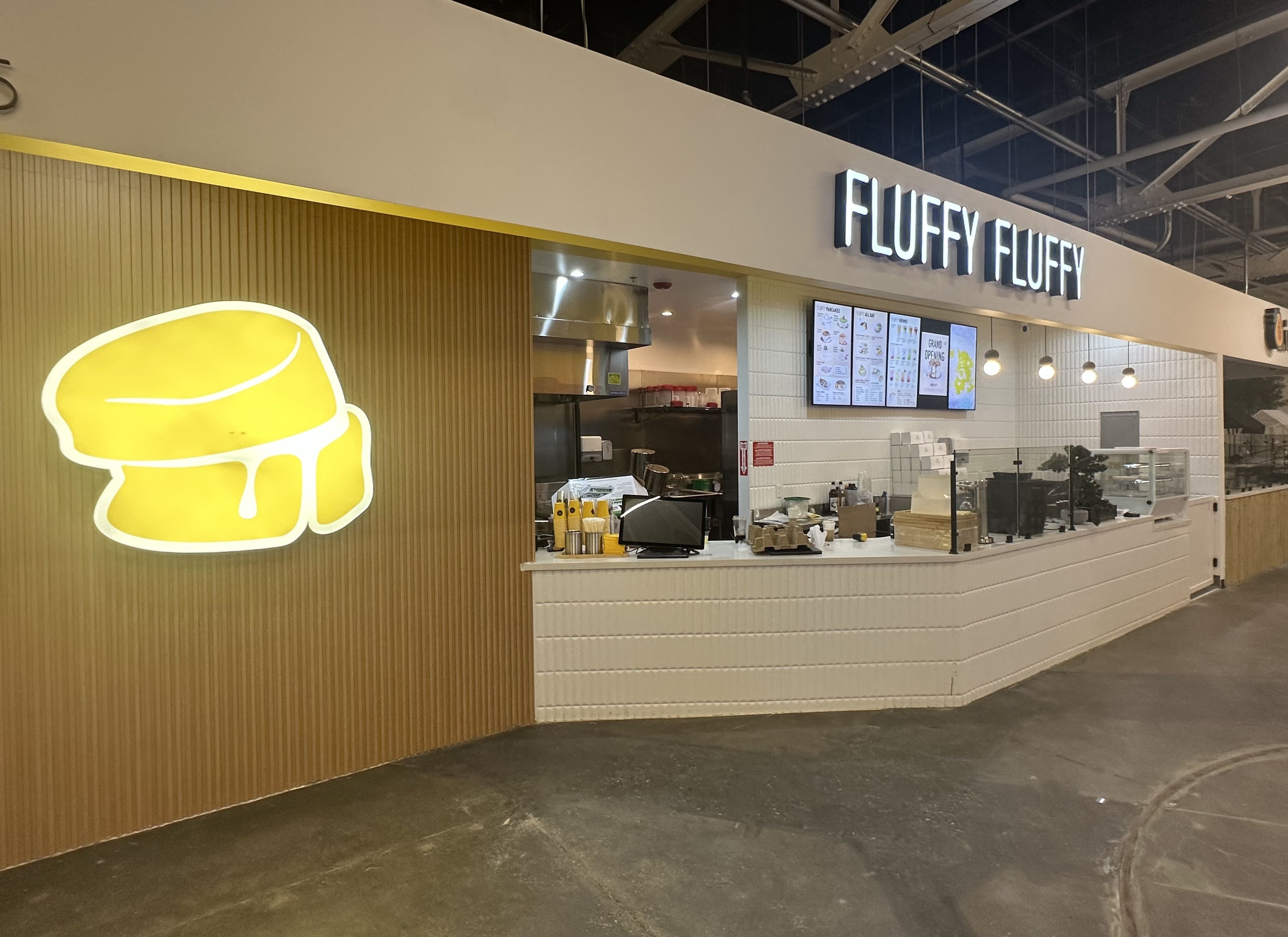 Inside a mall dessert shop, a white counter with tiled wall and wooden panel. A bright yellow neon lemon sign sits left; overhead, the sign "FLUFFY FLUFFY" and menu boards are visible.